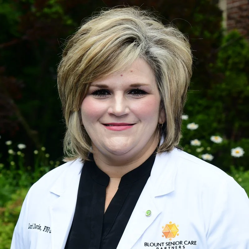 A woman with blonde hair wearing a white medical coat outdoors with greenery and flowers in the background.