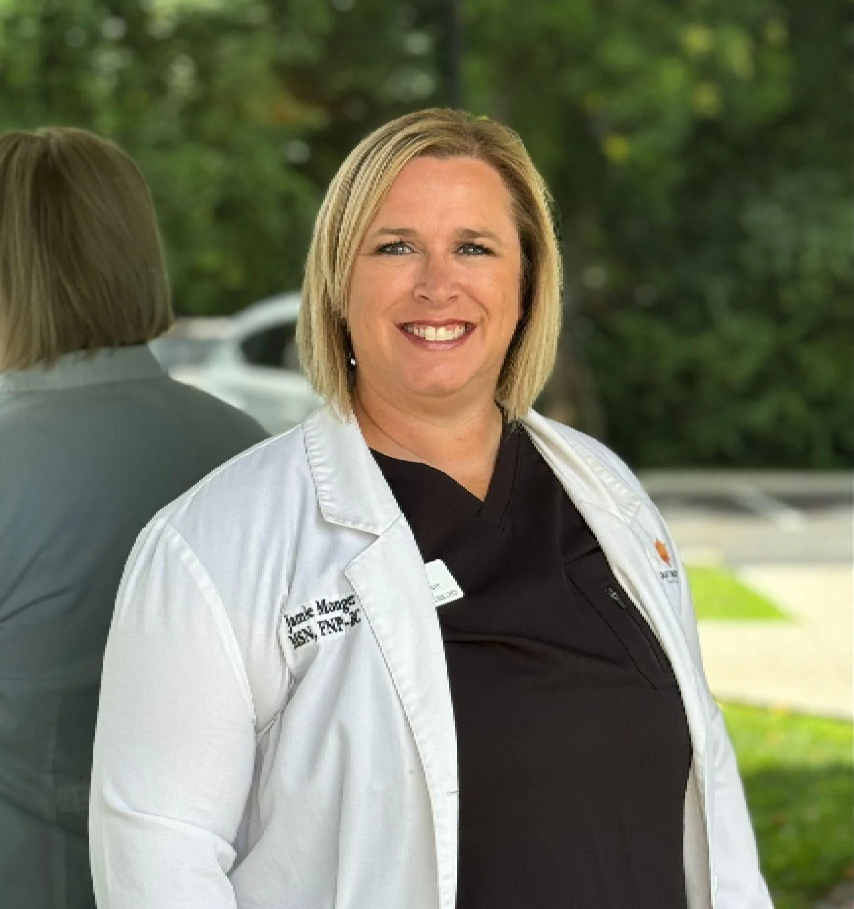 A smiling woman wearing a white medical coat standing outdoors with a green, leafy background and her reflection visible in a mirror behind her.