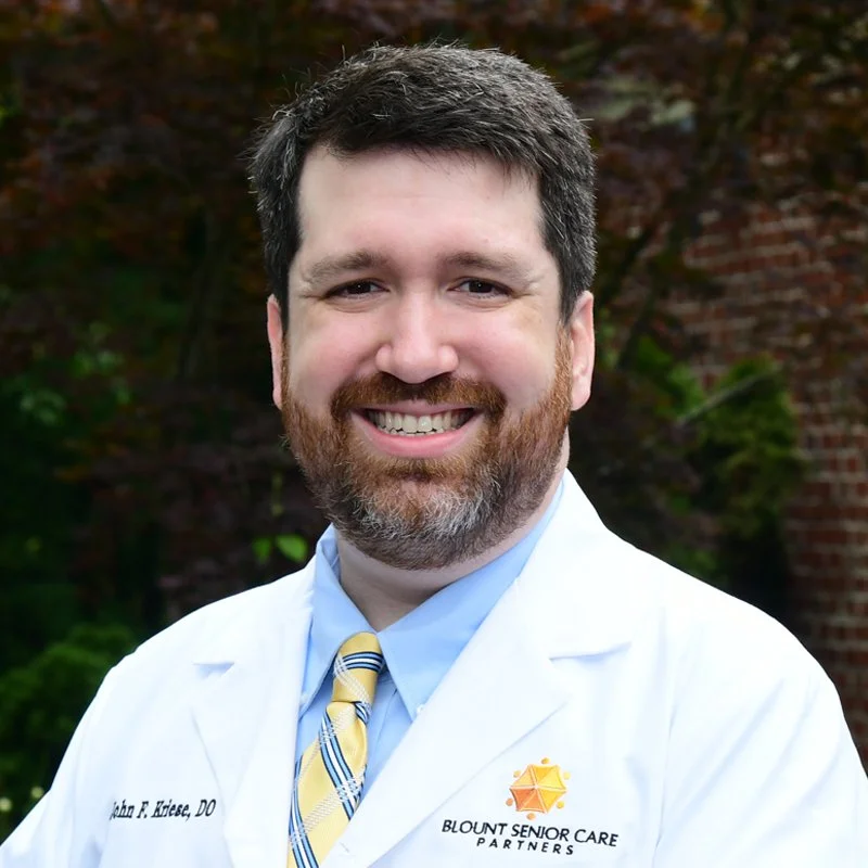 A smiling man with dark hair, beard, and mustache wearing a white medical coat outdoors with greenery and a brick building in the background.