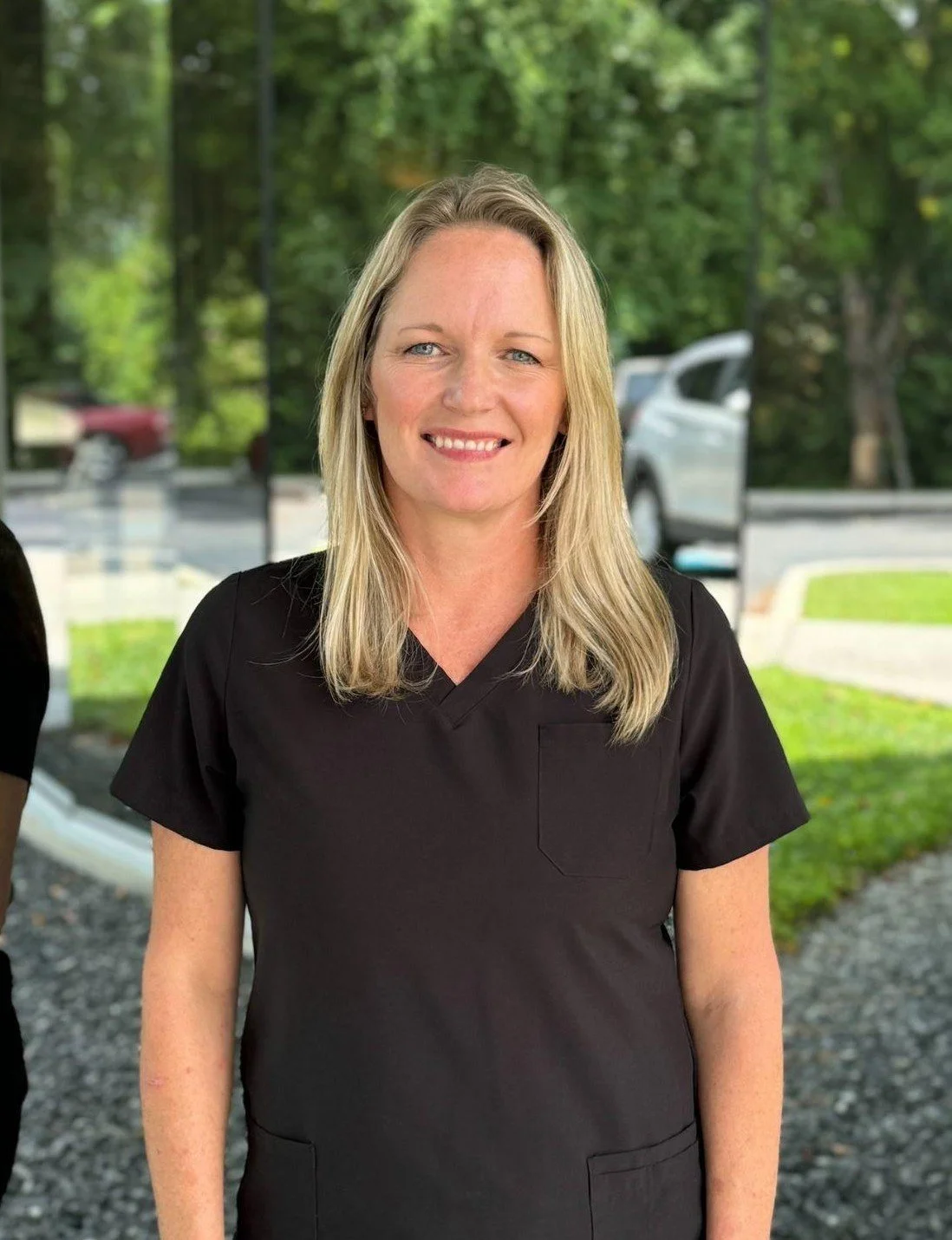 A woman with blonde hair smiling outdoors, wearing black scrubs, with trees and parked cars in the background.