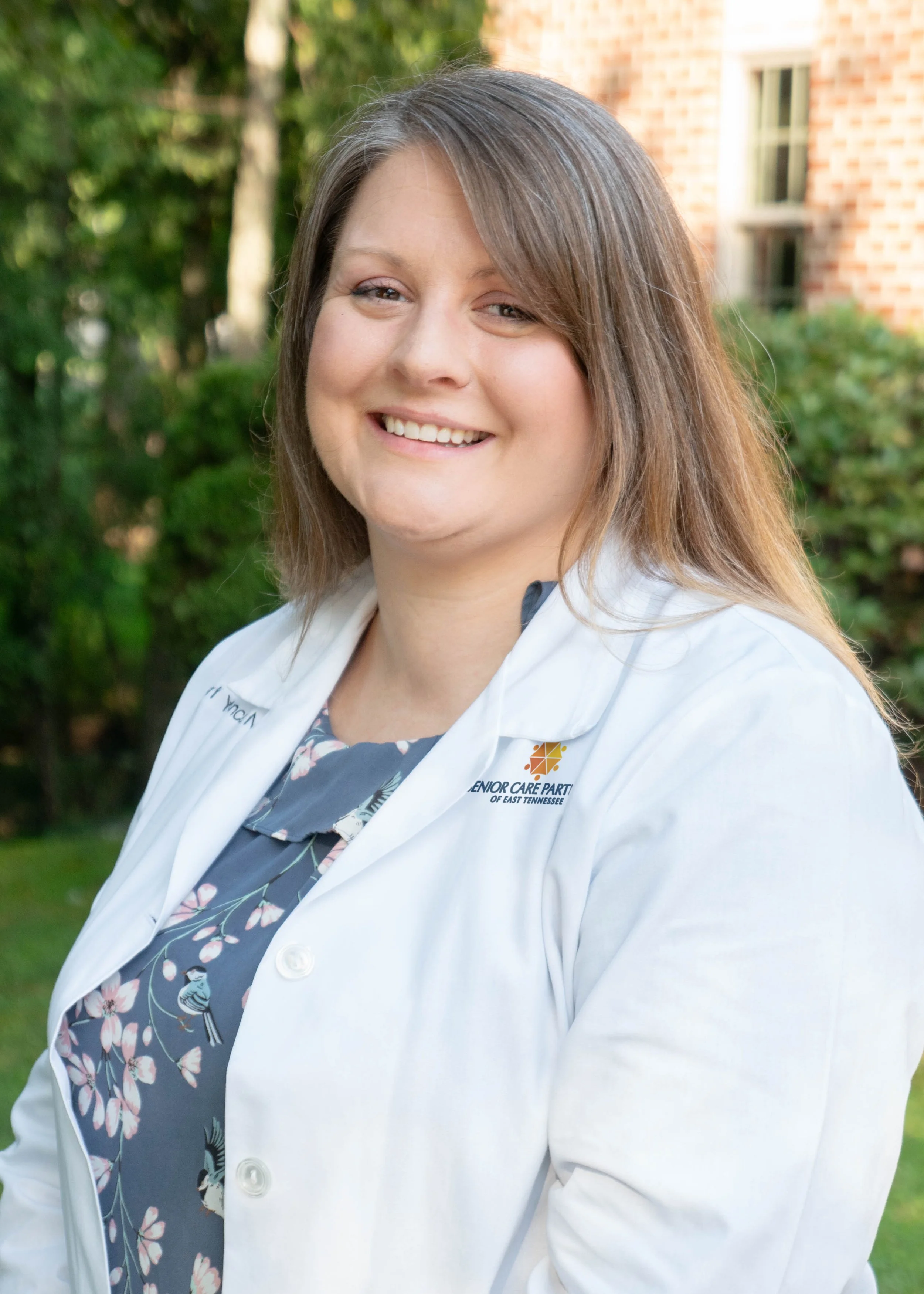 A woman with long light brown hair smiling outside, wearing a white coat with a logo that reads 'Senior Care Partners of East Tennessee' and a navy blue floral top, standing in front of greenery and a brick building.