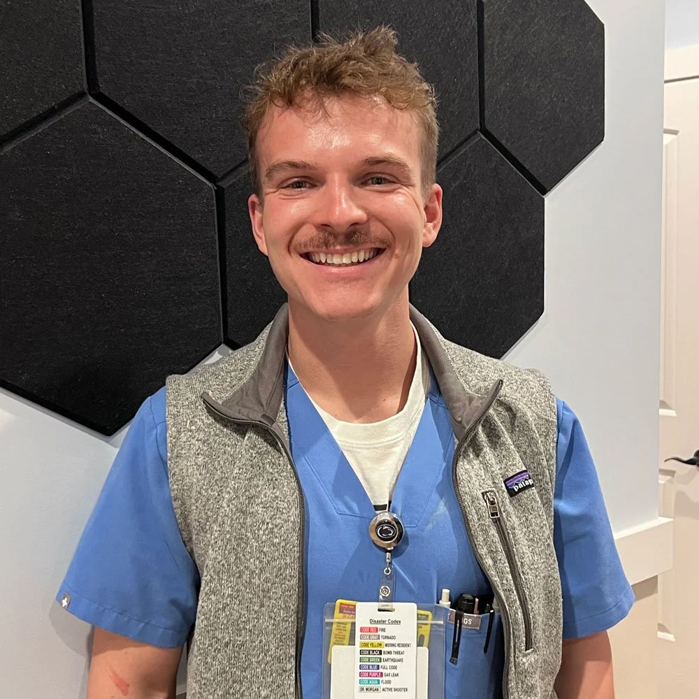 A smiling young man with light brown hair, wearing blue medical scrubs, a gray vest, and a YMCA badge, standing in front of a black honeycomb acoustic panel on a light-colored wall.