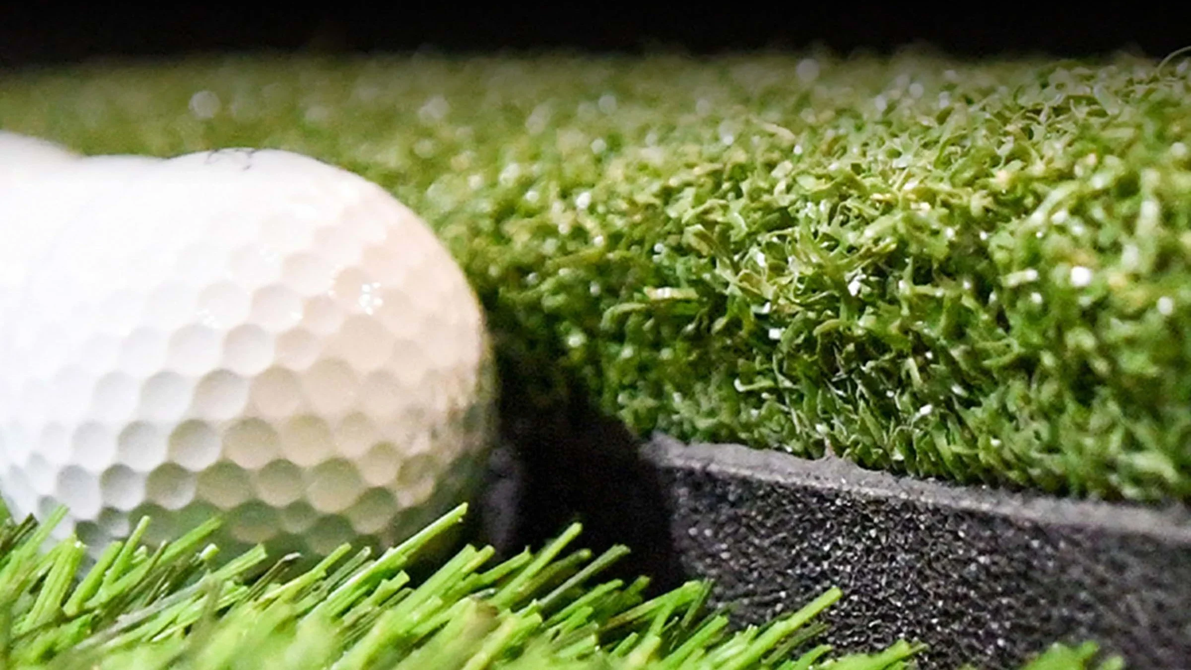 Close-up of a golf ball on Fiberbuilt artificial green grass with a section of a black tee and a green artificial turf in the background.