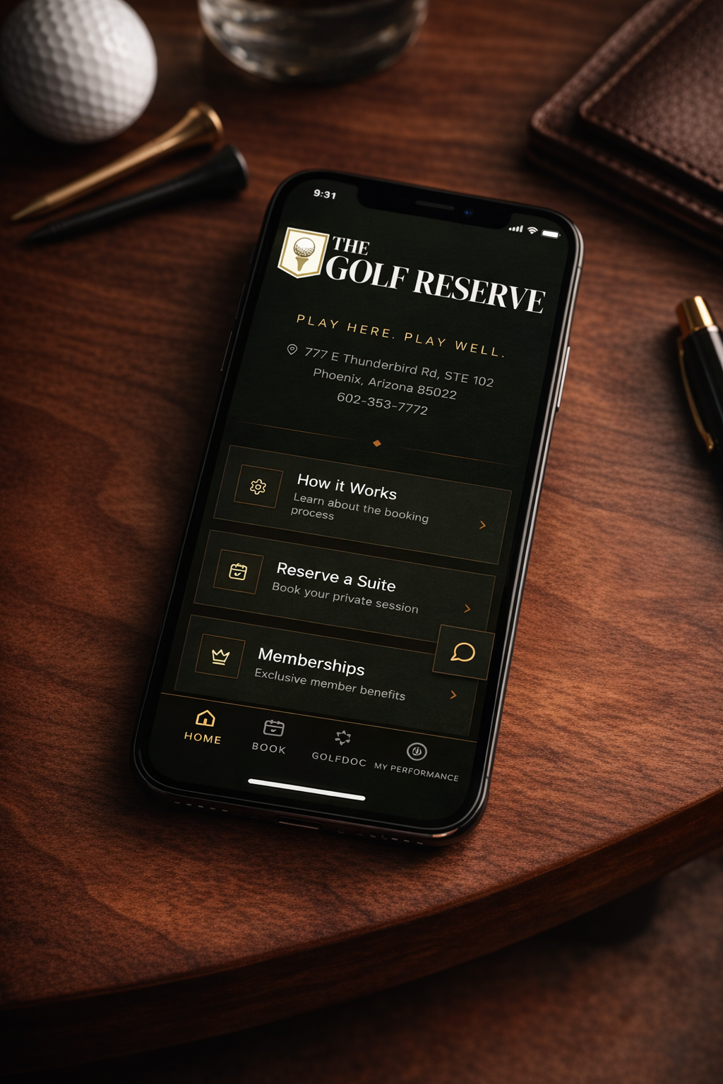 Smartphone displaying The Golf Reserve mobile app on a wooden desk with golf balls, pens, a notebook, and a glass of water.