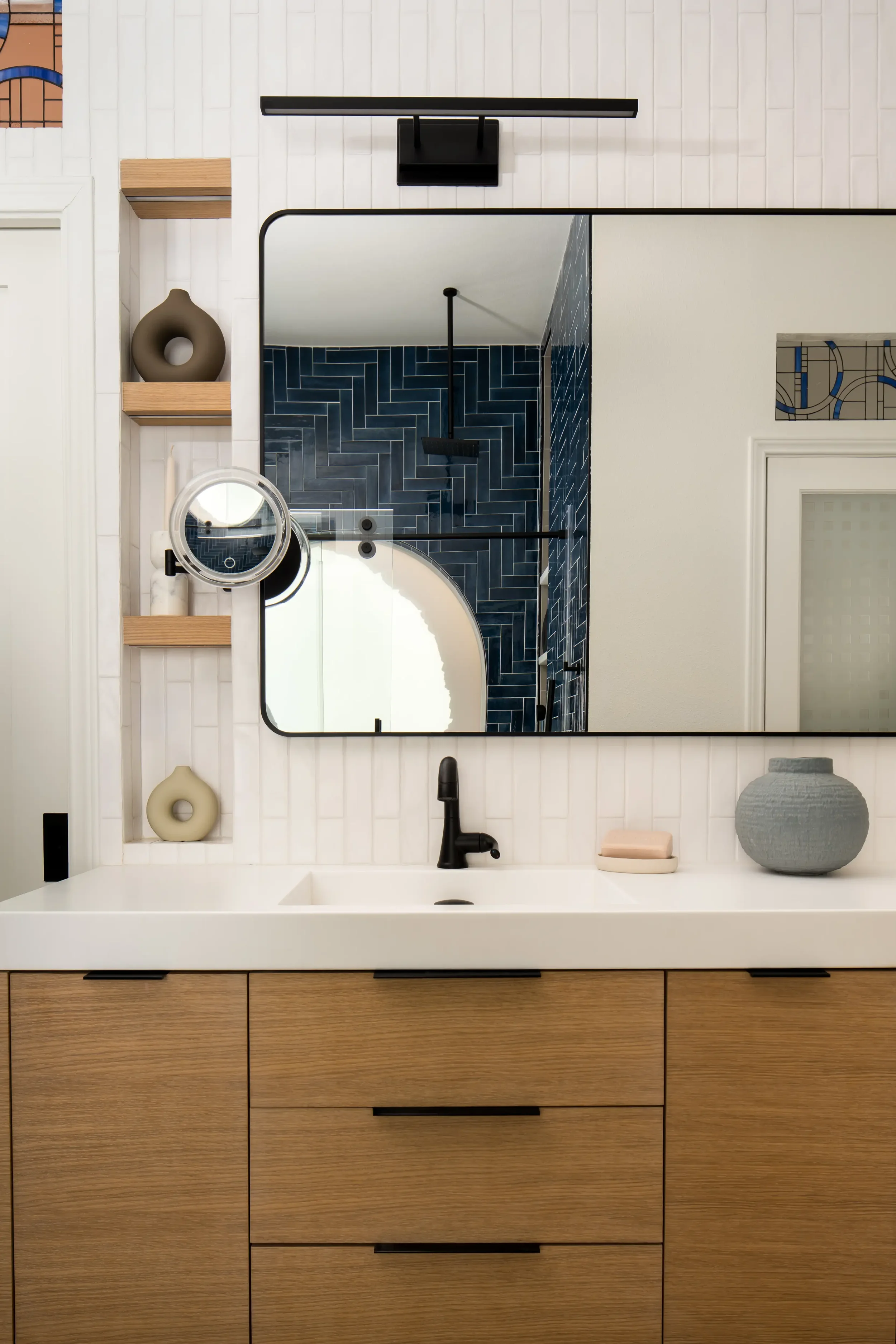 Modern bathroom vanity with a white countertop, black faucet, round mirror, beige soap dish, gray textured vase, and decorative objects. A large wall mirror reflects dark blue subway tile behind a black showerhead. Located in Dallas, TX