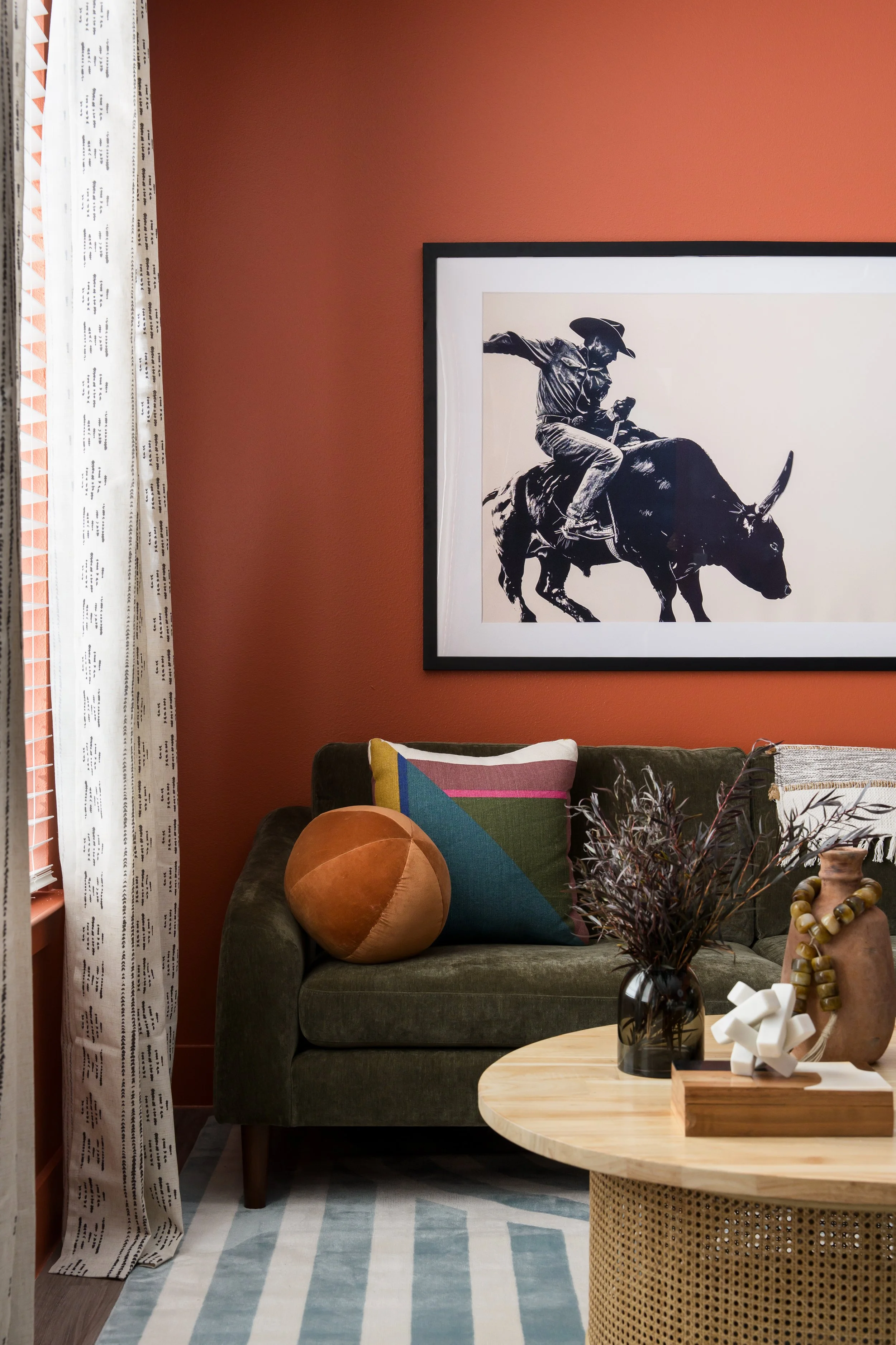 Living room with a dark green sofa, colorful throw pillow, round peach pillow, abstract rug, wooden coffee table with decorative items, framed black-and-white cowboy riding a bull artwork, red wall, window with patterned curtains.