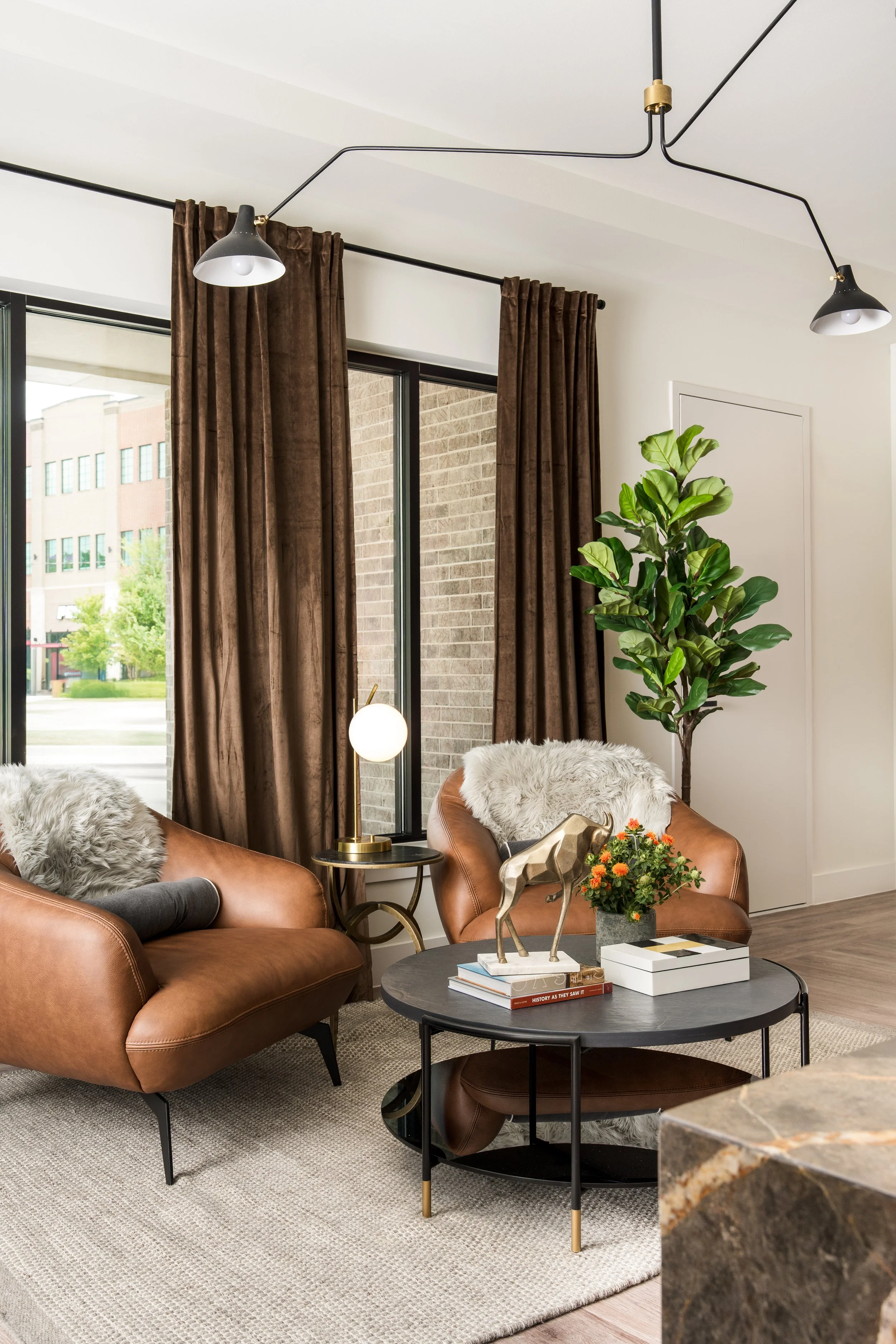 Living room corner with two leather armchairs, side table, indoor plant, and decor items including a table lamp, a golden deer sculpture, and books on a round coffee table. Large windows with brown curtains and a view of a building and greenery outsi