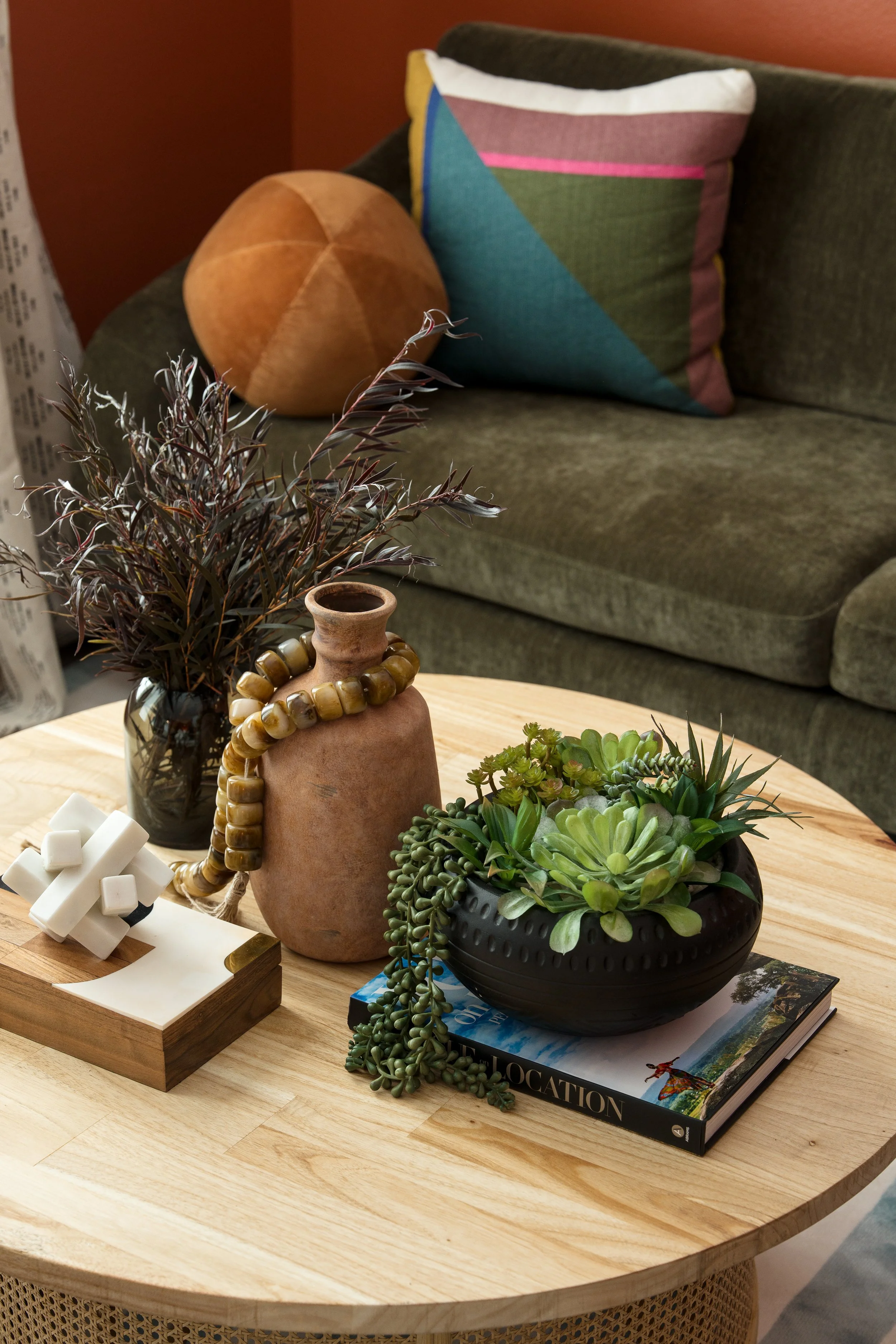 Close-up of a wooden table with decorative items, including potted plants, a tan pottery vase with a beaded necklace, a small wooden sculpture, and a book titled 'Location', with a dark brown sofa with pillows and a wall in the background.