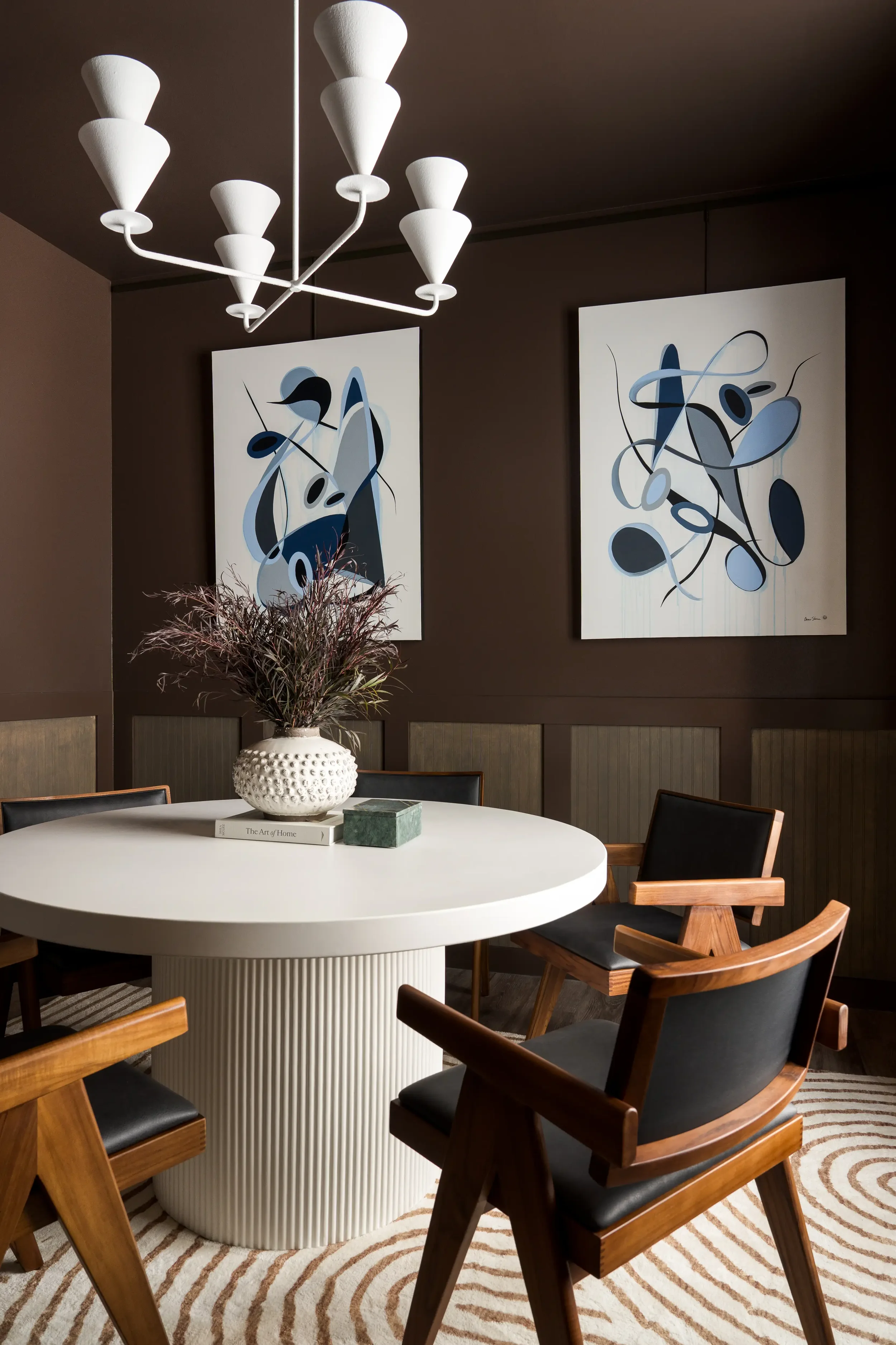 Interior of a modern dining room with a white round table, black and wooden chairs, a white textured vase with pink and green plants, abstract art paintings in shades of blue and black, a contemporary white chandelier, and a striped beige and brown r