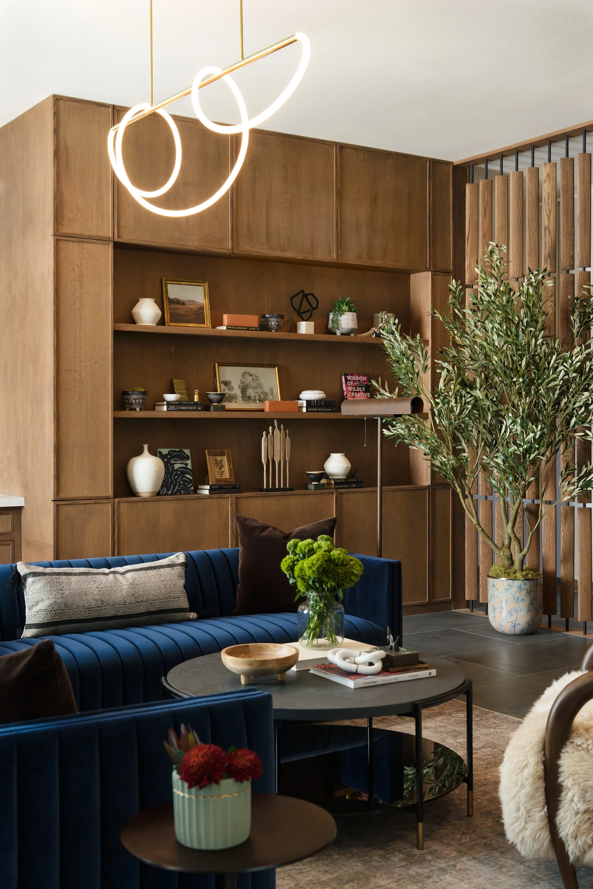 Lounge at The Gallery in Frisco, Texas, with a navy channel-tufted sectional, warm wood cabinetry, an olive tree, and a sculptural ring pendant, designed by Artbuckle & Co.