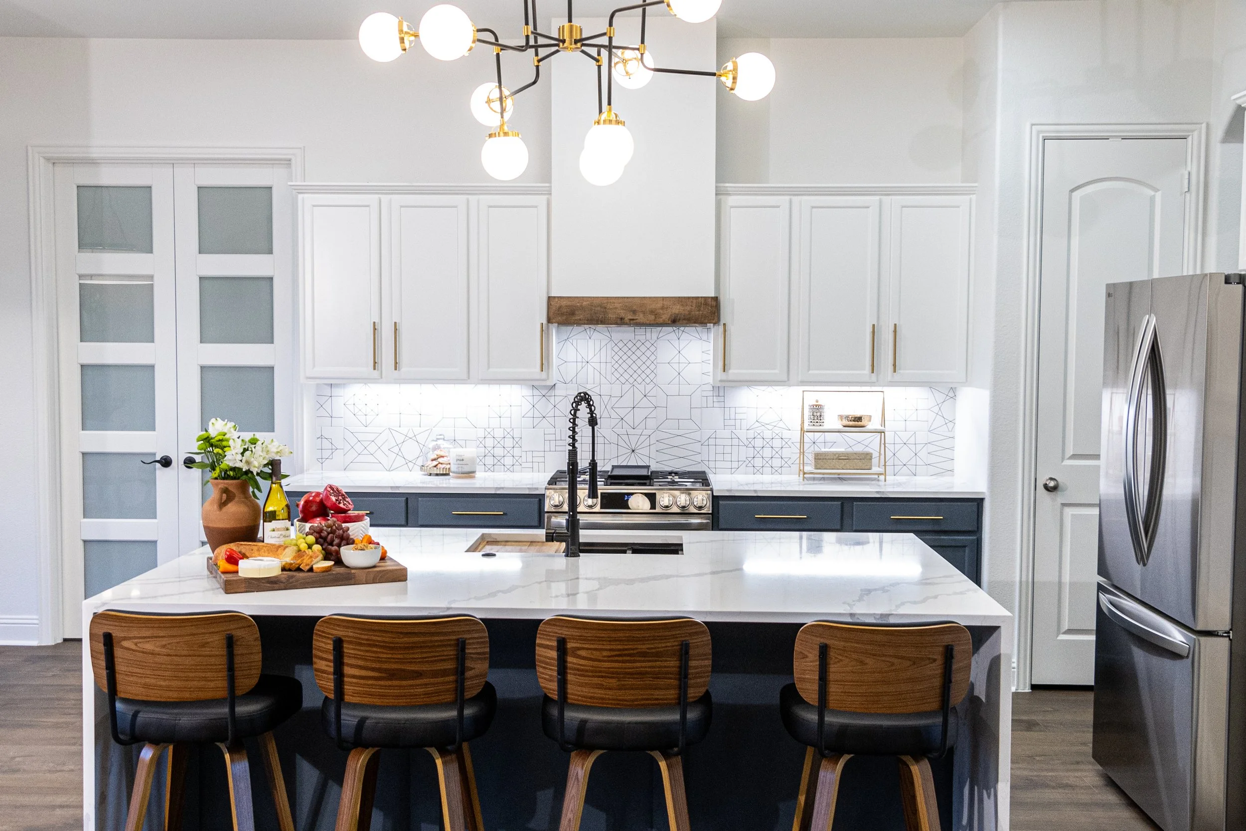 A modern kitchen with white cabinets, a blue island with a white marble countertop, and a black and gold chandelier above. The kitchen features a stainless steel refrigerator, a black faucet, and a breakfast bar with wooden chairs. There is a vase wi
