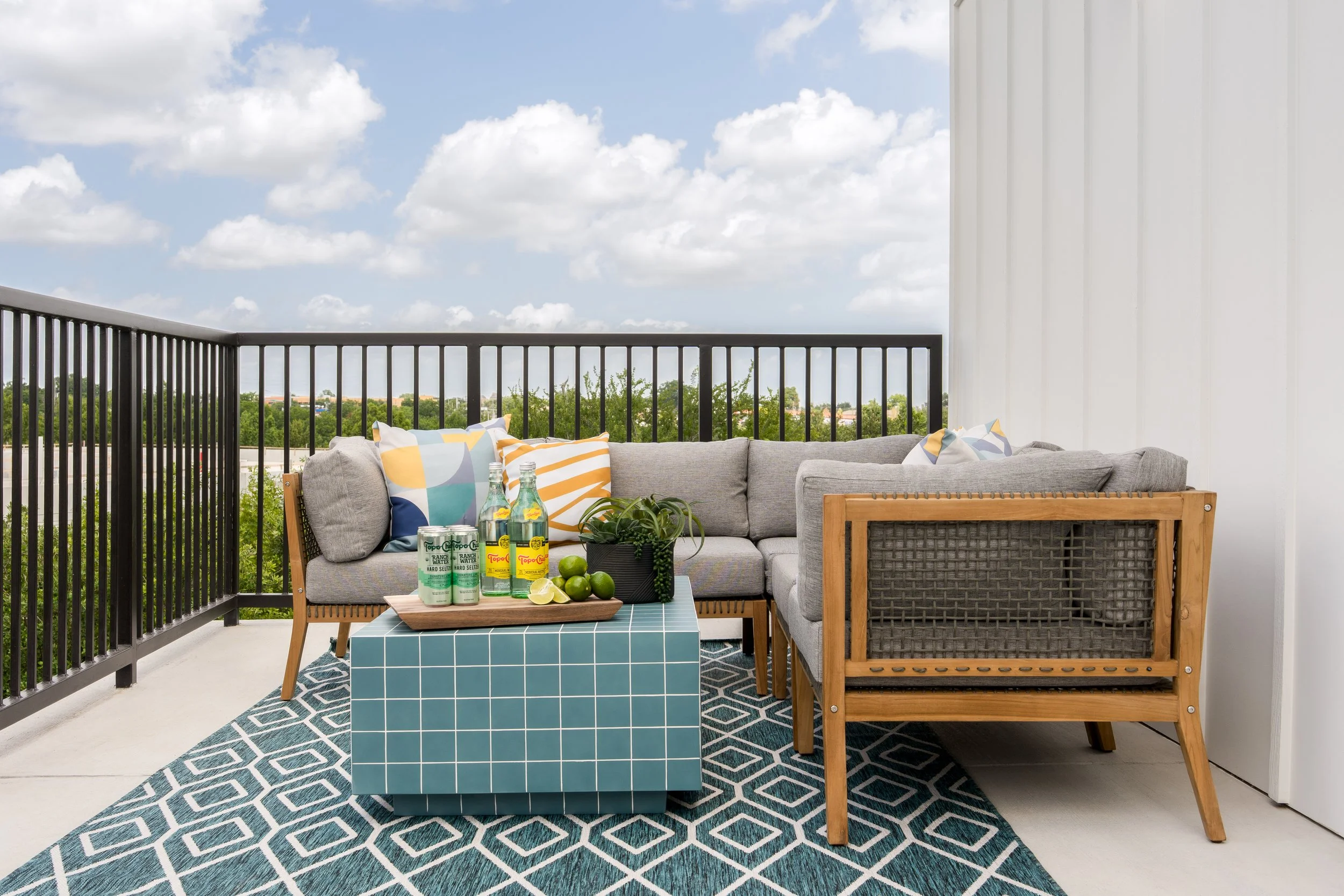Balcony outdoor seating area with gray cushioned sofa with colorful pillows, a blue tiled coffee table with drinks and lime, a patterned outdoor rug, black metal railing, white wall, and a view of green trees and blue sky with clouds.