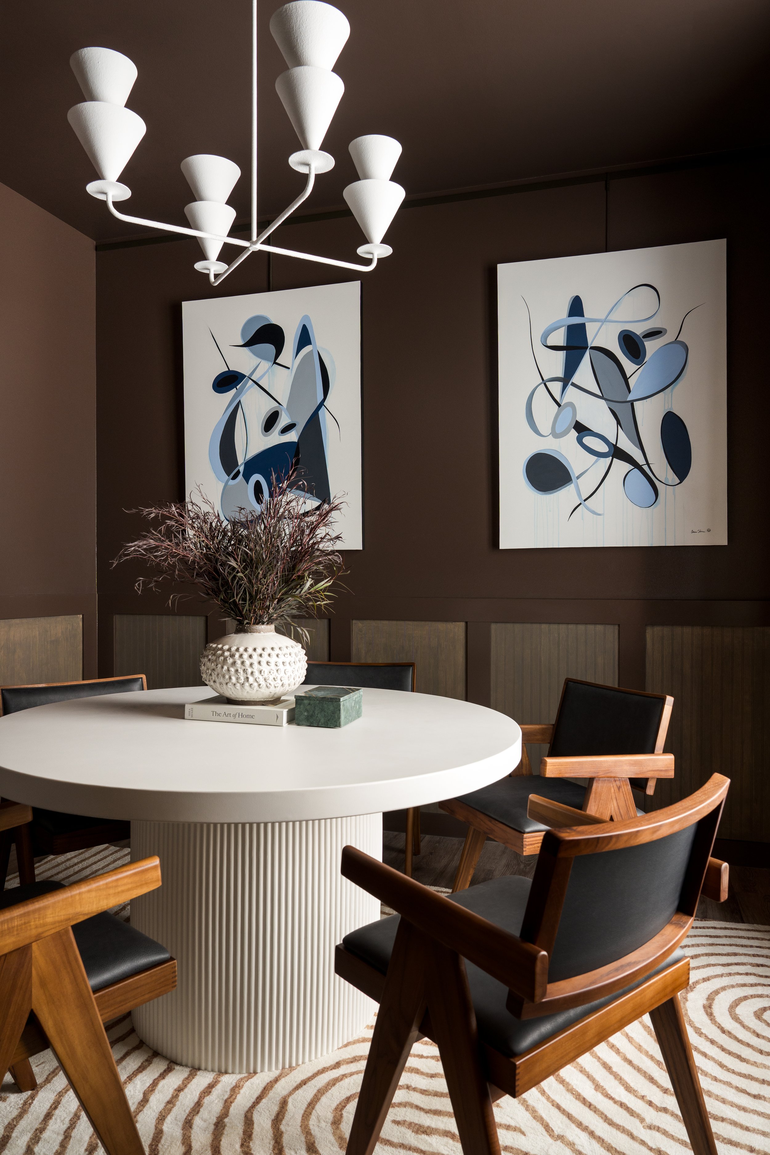 Modern dining room with white round table, wooden chairs with black cushions, abstract wall art, a floral arrangement in a textured white vase, and a geometric patterned rug.