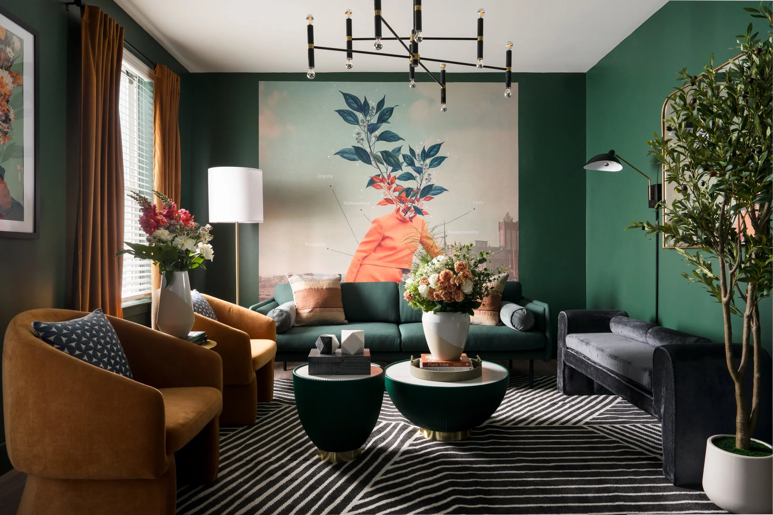 A modern living room with green walls, featuring a large wall art with plant illustration, a mix of velvet and fabric sofas, a black and gold chandelier, and decorative plants and flowers.