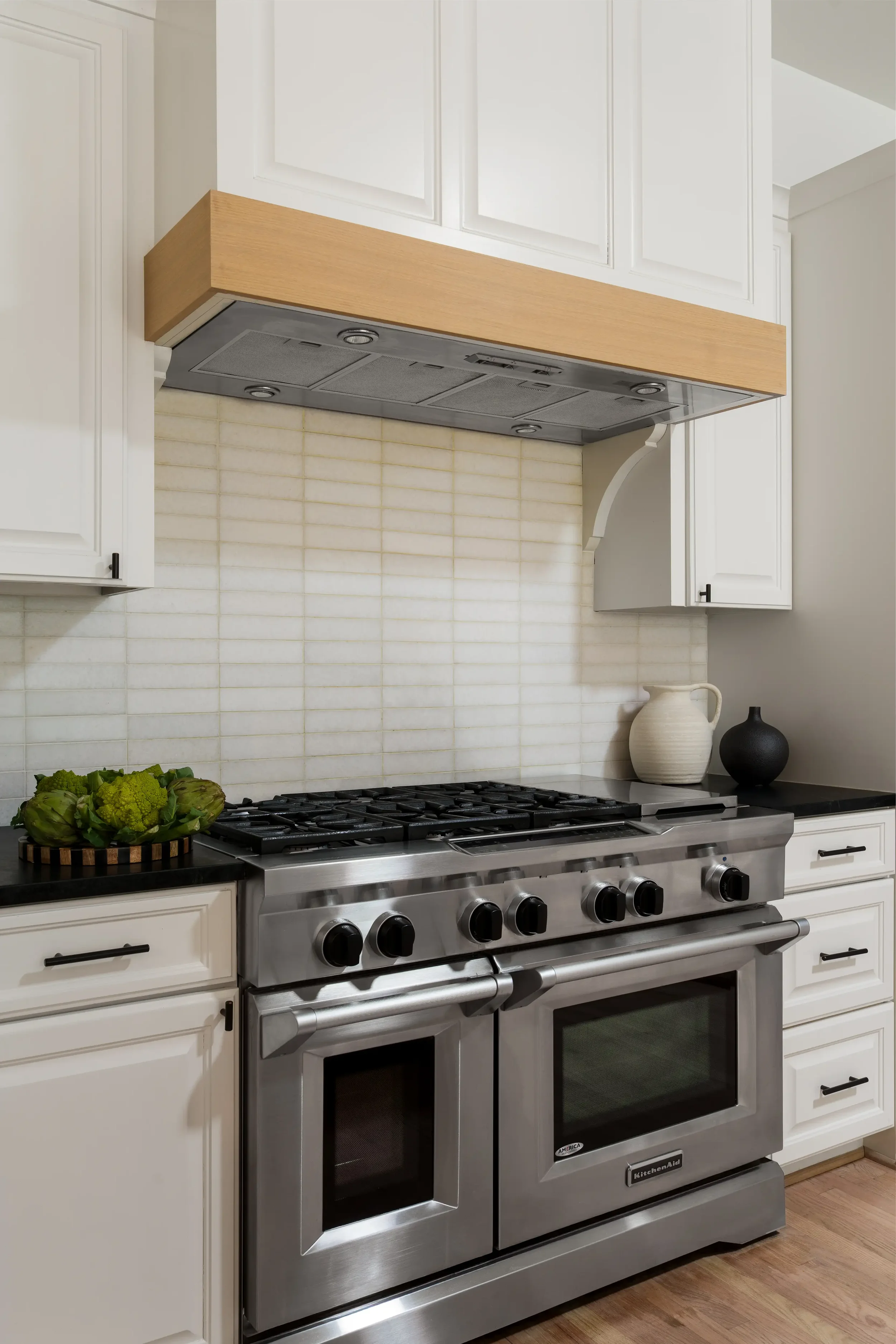 Kitchen range wall in a Lakewood Hills home in Dallas, Texas, with white cabinetry, a light wood range hood, a stainless steel range, and a pale tile backsplash, designed by Artbuckle & Co.