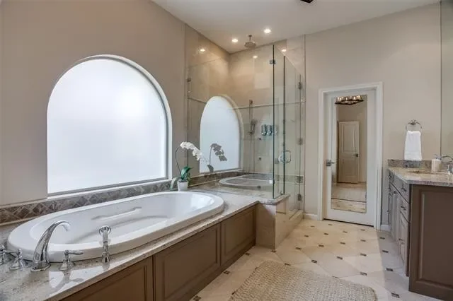 Before: the primary bathroom in a Lakewood Hills home in Dallas, Texas, with beige walls, a drop-in soaking tub with a stone surround, a corner glass shower, and arched frosted windows, prior to the Artbuckle & Co. renovation.