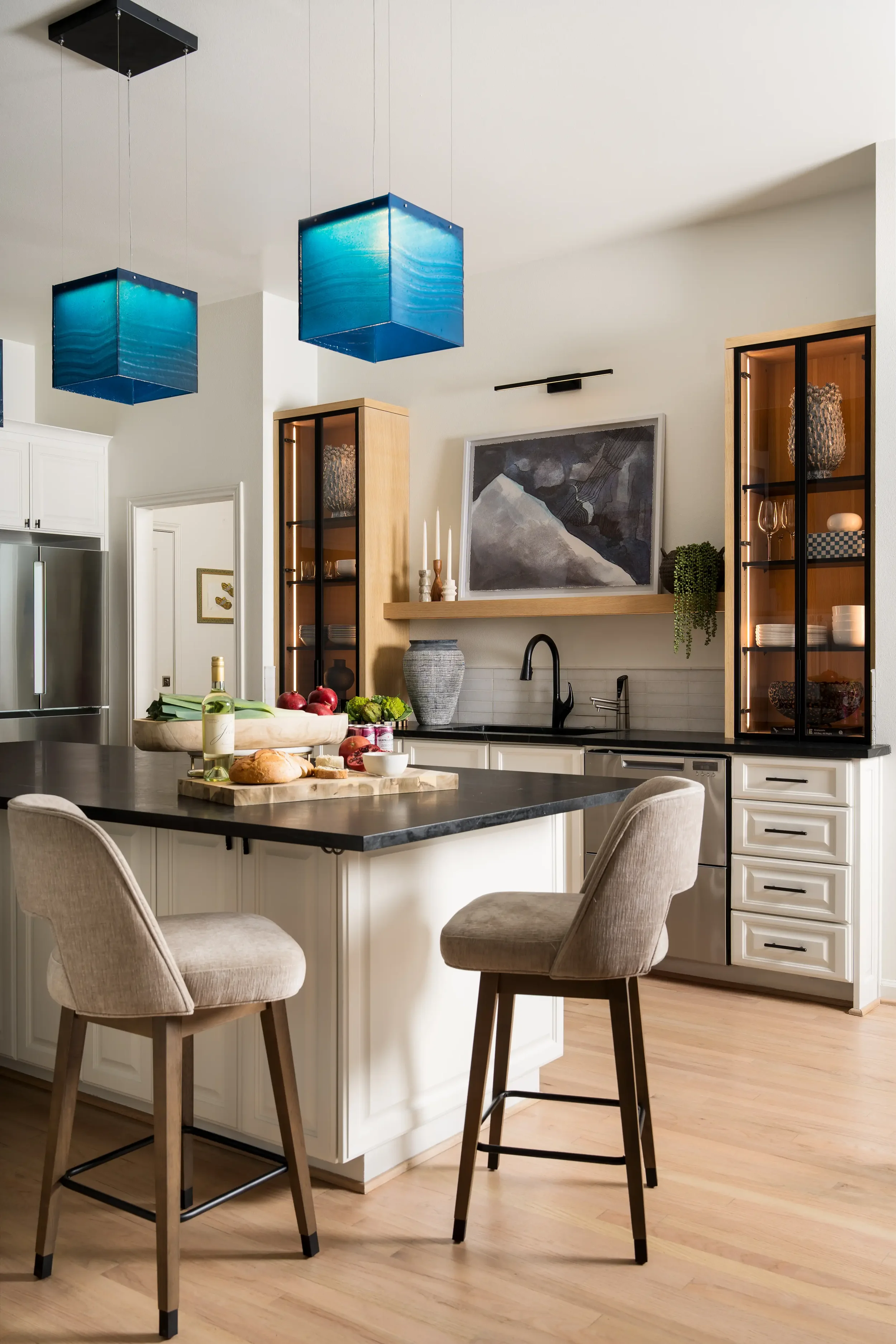 Modern kitchen with white cabinetry, black countertop, and two beige chairs at the island. There are two blue pendant lights hanging from the ceiling, and decorative items on the shelves and counter. Fresh produce and a bottle of wine are on the isla