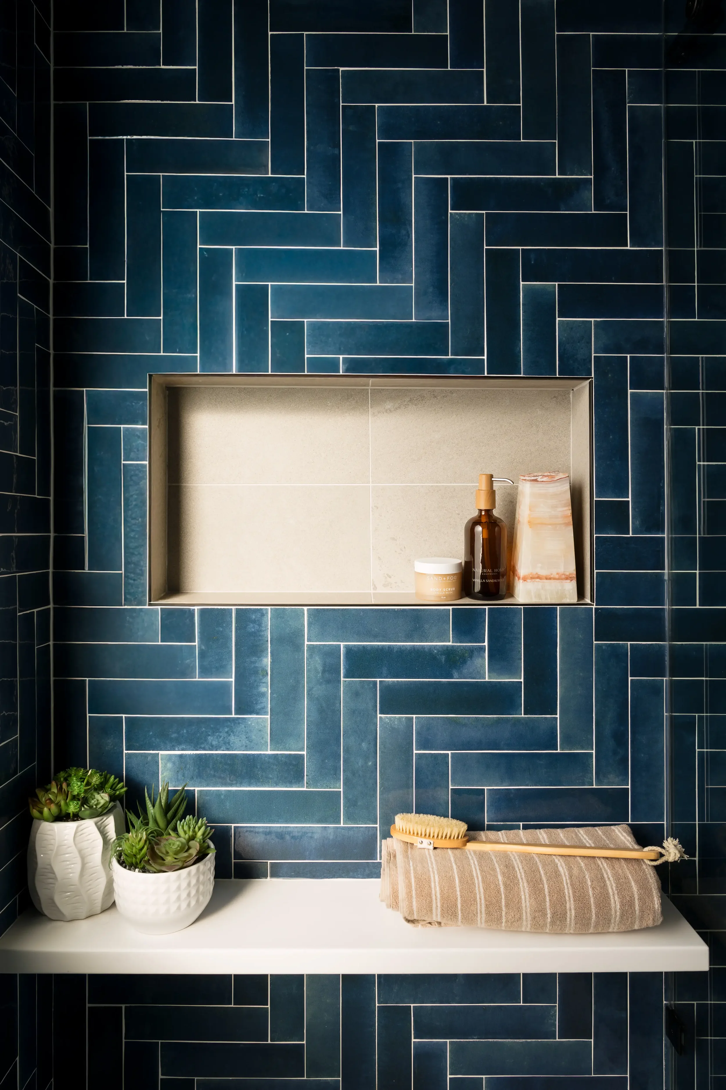 Interior bathroom shelf with potted succulents, skincare products, a rolled towel, and a brush, against dark blue tiled wall with geometric pattern. Located in Dallas, TX