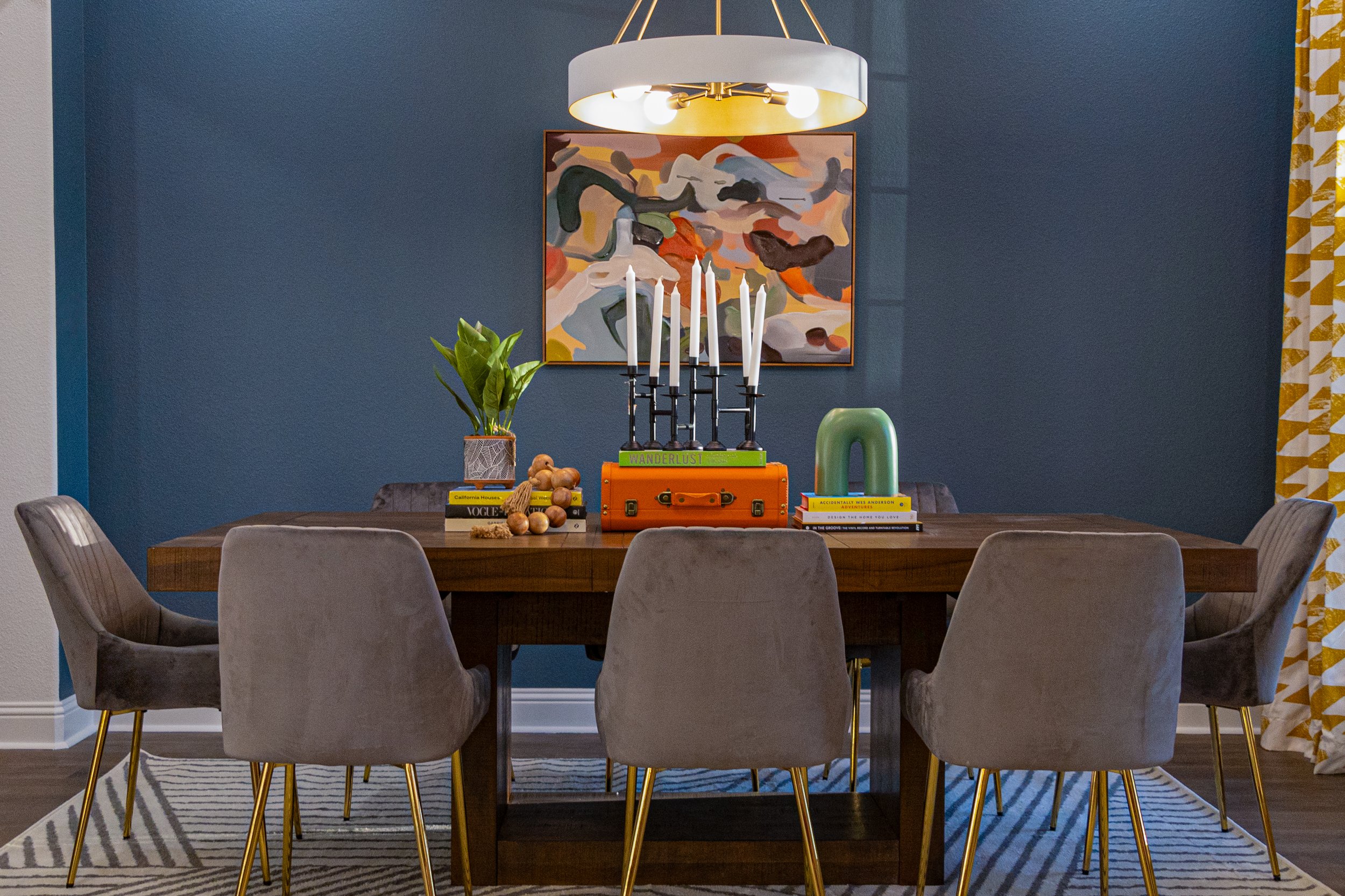 Dining room with a wooden table, six gray upholstered chairs with gold legs, a white and gold chandelier, a colorful abstract painting on a blue wall, a potted plant, candlesticks, and a stack of books on the table, with patterned curtains on the rig