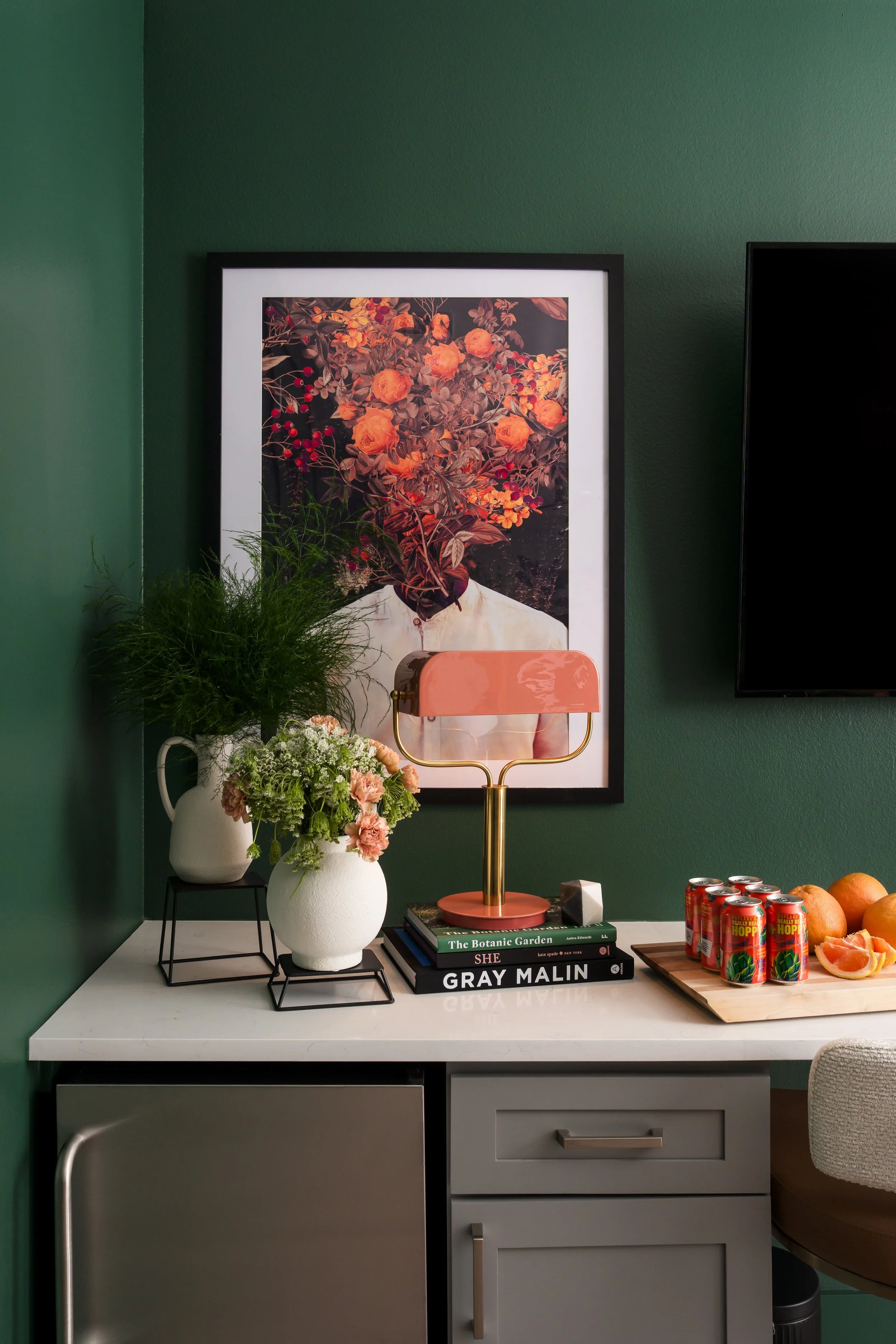 A white cabinet with two drawers and a door, topped with a white marble surface, is against a green wall. On the surface, there are two white vases with pink and green flowers, stacked on black metal stands. Above the cabinet, a large framed poster o