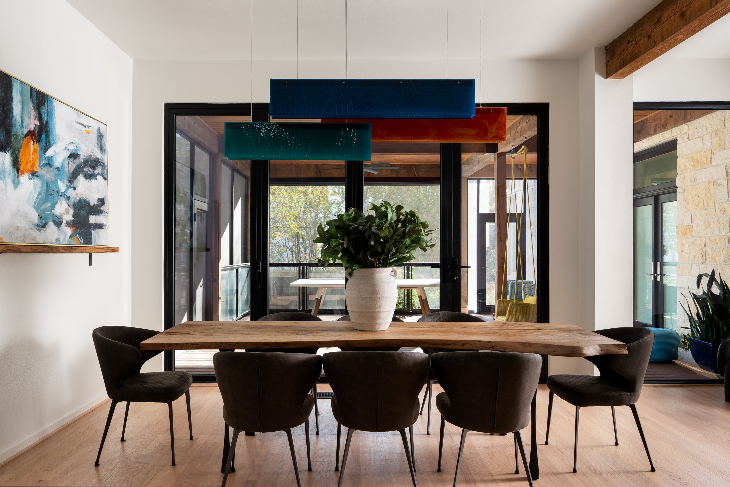 Modern dining room with a wooden table, black chairs, a large potted plant, and abstract art on the white wall. Large glass doors lead to an outdoor patio with wooden beams and greenery.
