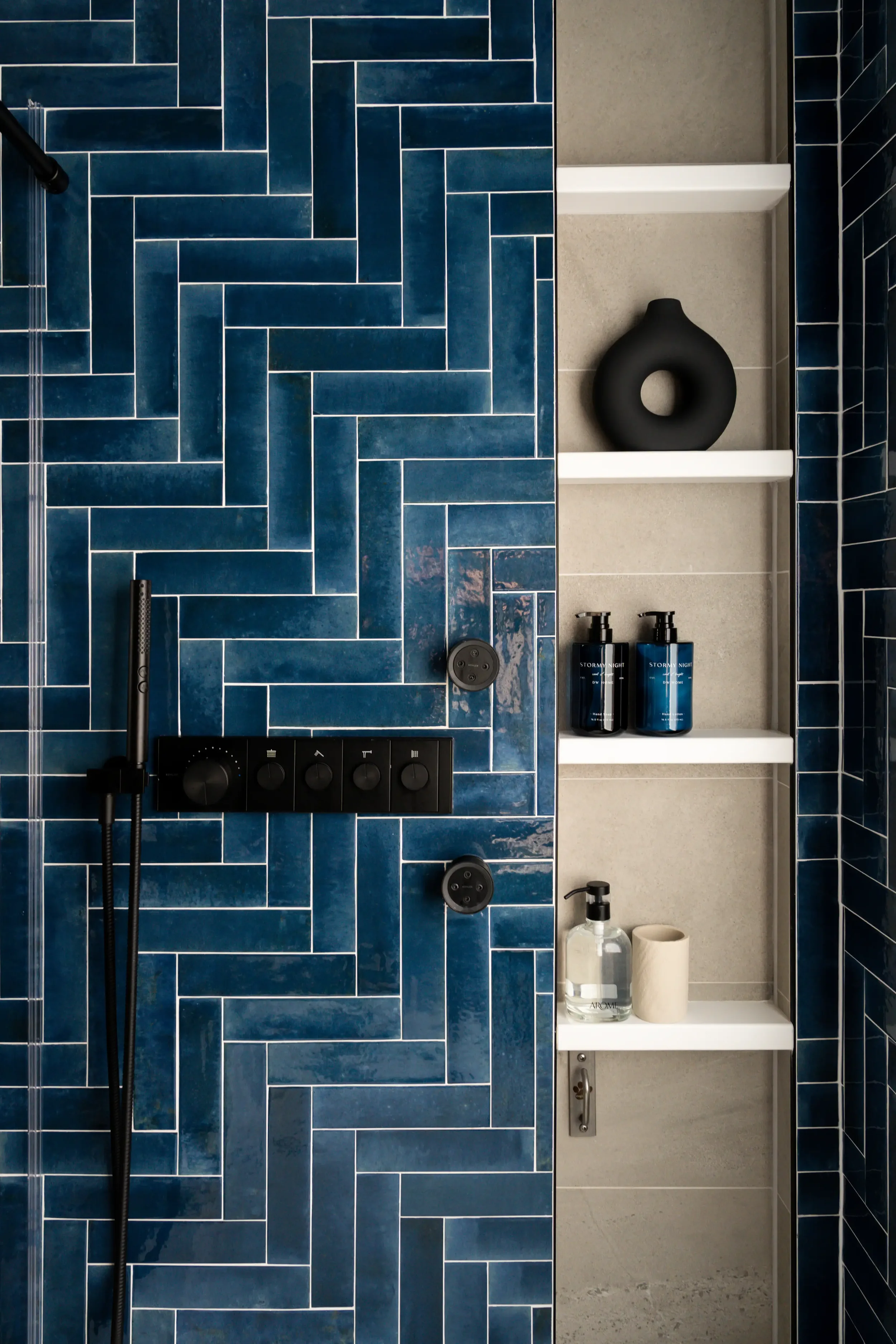 A modern bathroom shower area with blue herringbone tiles, black shower fixtures, a white shelf with a black decorative vase, bottles of soap, a toilet paper roll, a clear bottle of liquid, and a soap dispenser. Located in Dallas, TX