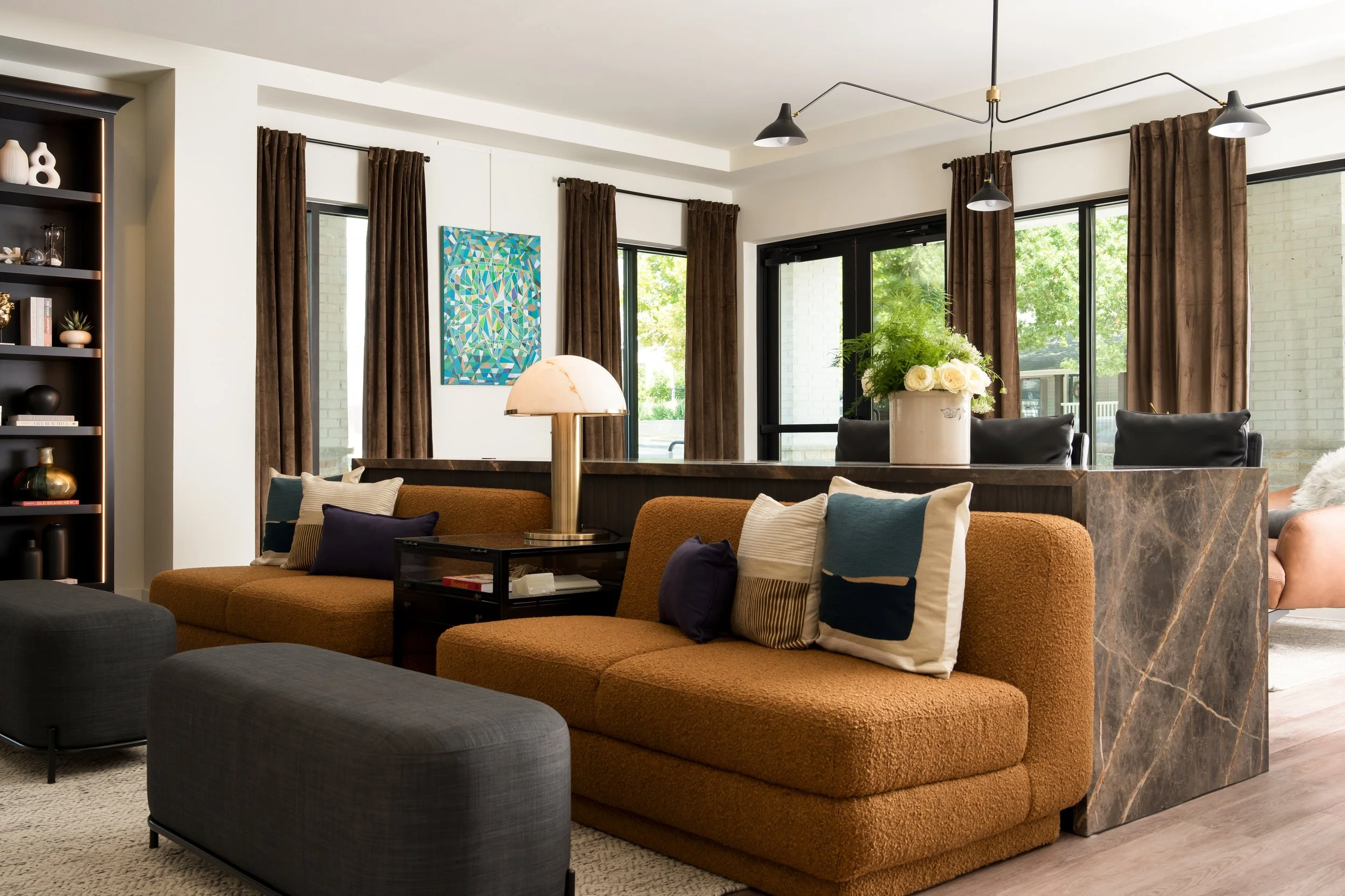 Living room with brown sofa, decorative pillows, a black ottoman, a marble-topped table with a large white flower vase, and open black-framed glass doors with brown curtains, overlooking a green outdoor area.
