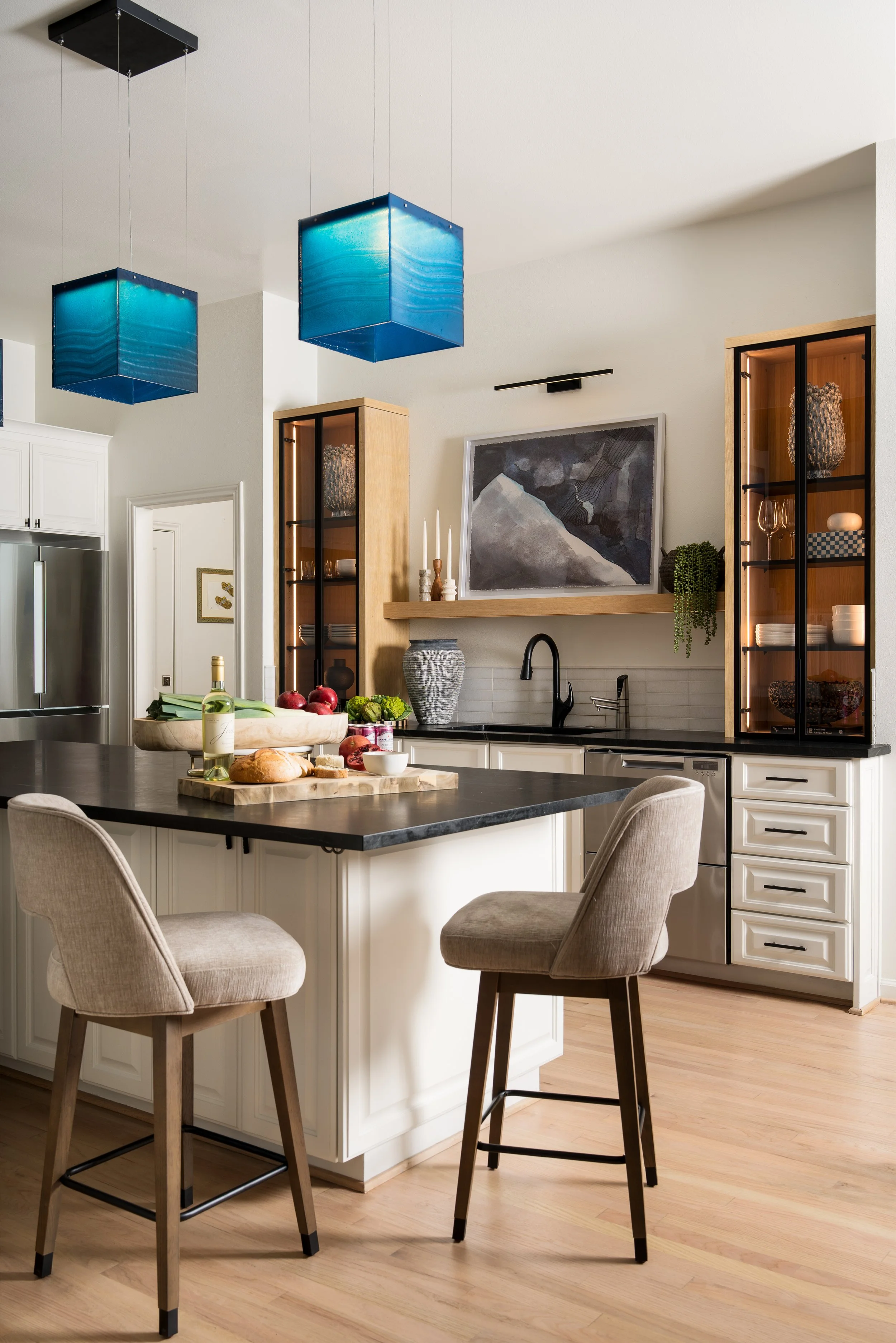 Modern kitchen with white cabinets, black countertops, and beige chairs. Blue pendant lights hang above the breakfast bar. Decor includes artwork, plants, and various kitchen items.