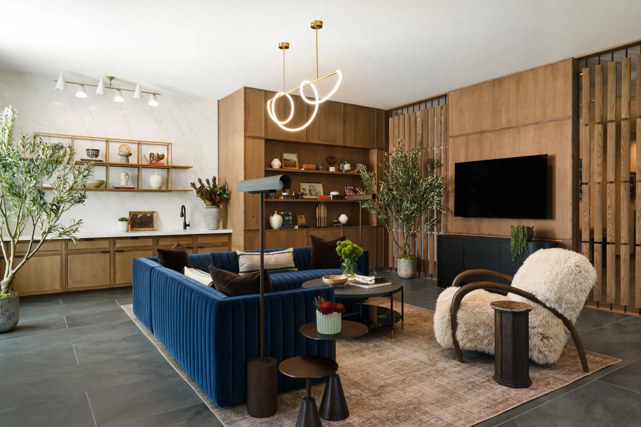 Angled view of The Gallery lounge in Frisco, Texas, with a navy tufted sectional, a looping pendant light, an olive tree, and a wood media wall, designed by Artbuckle & Co.