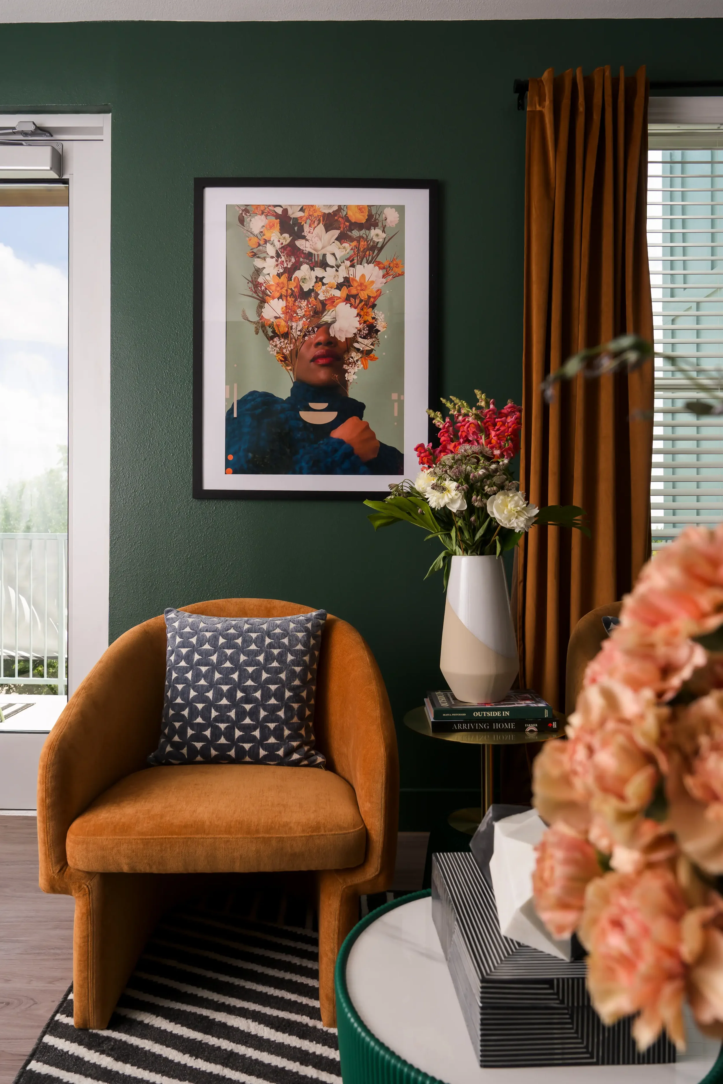 A cozy living room corner with an orange armchair, a decorative pillow, a small side table with a striped box, a vase of pink and white flowers, and a framed floral portrait on a green wall, next to a window with golden curtains. Located in Dallas, T