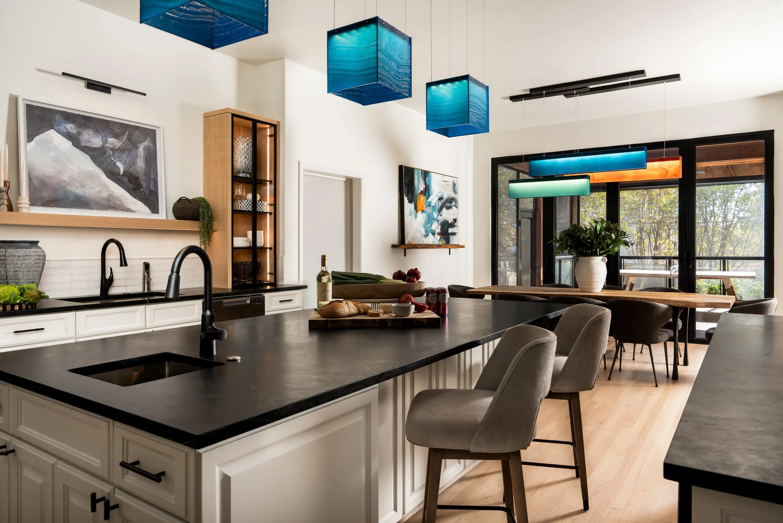 Modern kitchen with black countertop island, white cabinets, gray upholstered chairs, and large windows with a view of trees, decorated with abstract paintings and colorful pendant lights.