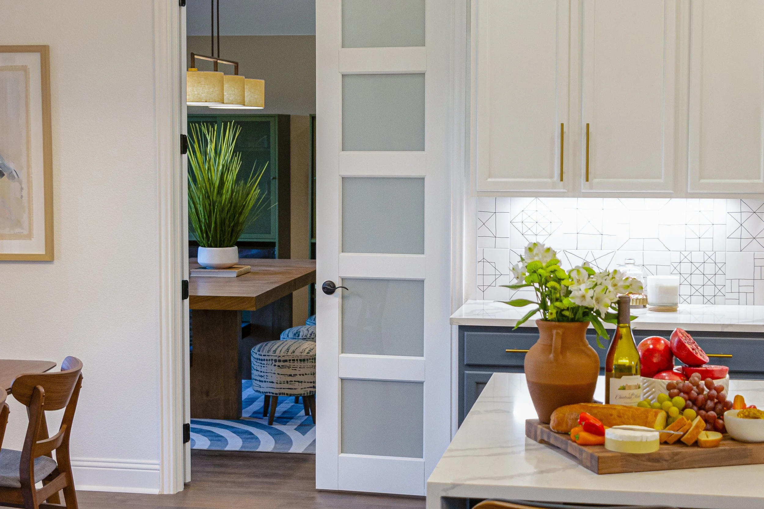 View of a modern kitchen with a white countertop, a vase with white flowers, and various fruits including grapes, watermelon, and cheese on a wooden board. In the background, there are white cabinets with gold handles, a white tiled backsplash, and a