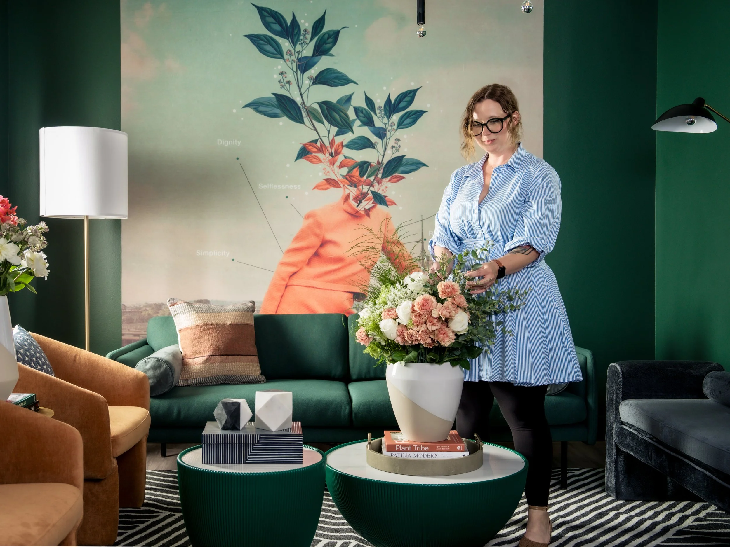 A woman in a blue striped dress arranging a large floral bouquet in a white vase on a white coffee table in a colorful living room with orange, green, and gray furniture and a wall mural of a person with plant-themed features.