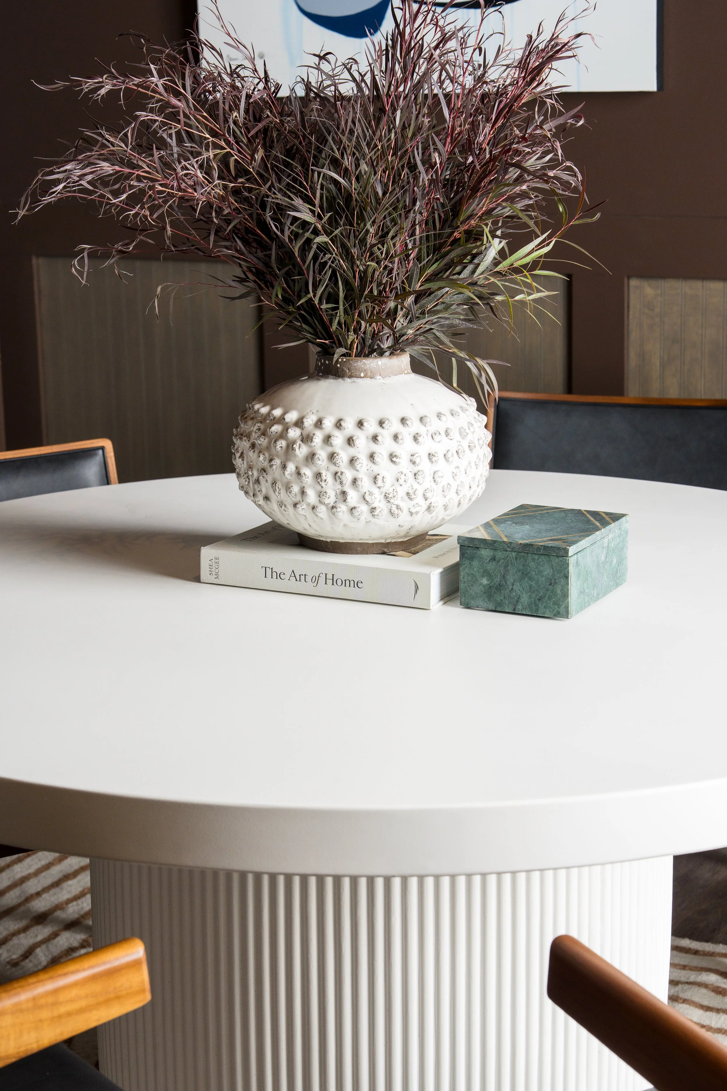 A round white table with a textured cylindrical base, a book titled 'The Art of Home' and a decorative box on top, and a large potted plant with long, purple and green leaves placed on the table, in a room with brown walls and a painting hanging abov