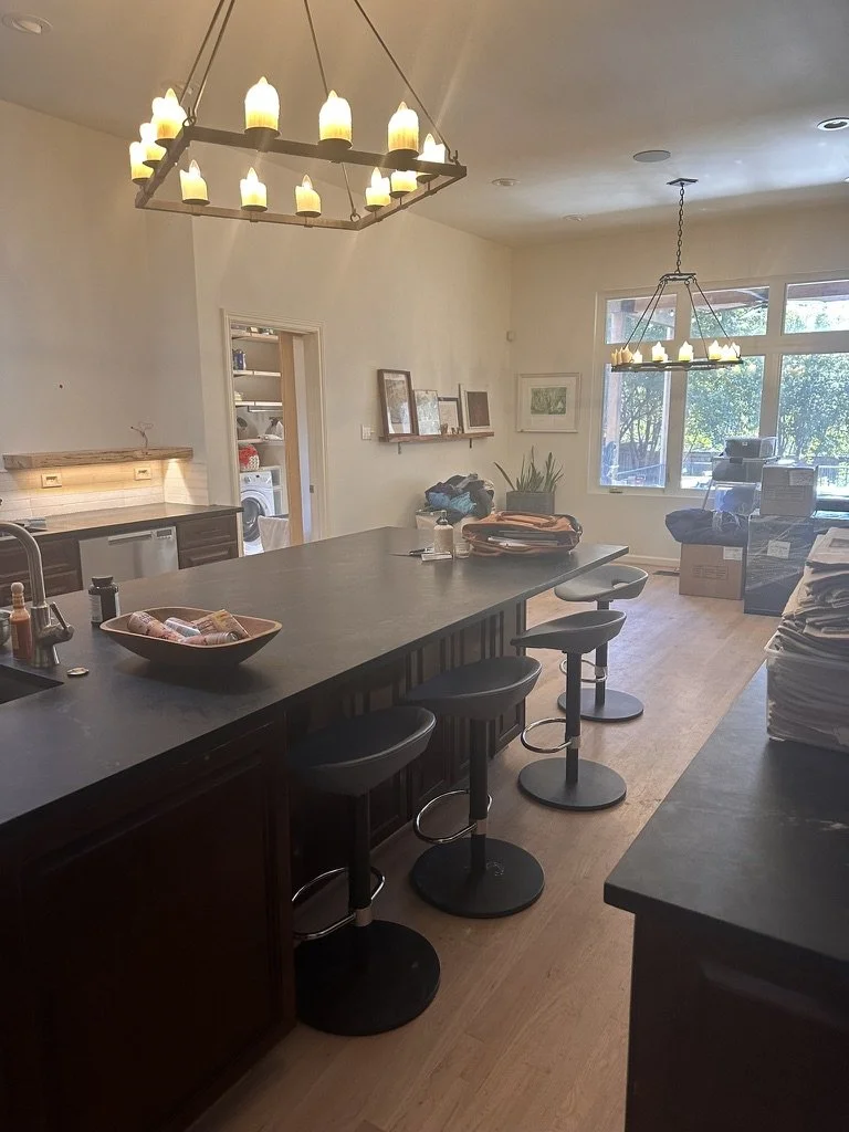 Before: the open kitchen and living area of a Lakewood Hills home in Dallas, Texas, with a long black-topped island, swivel bar stools, and candle-style chandeliers, prior to the Artbuckle & Co. renovation.