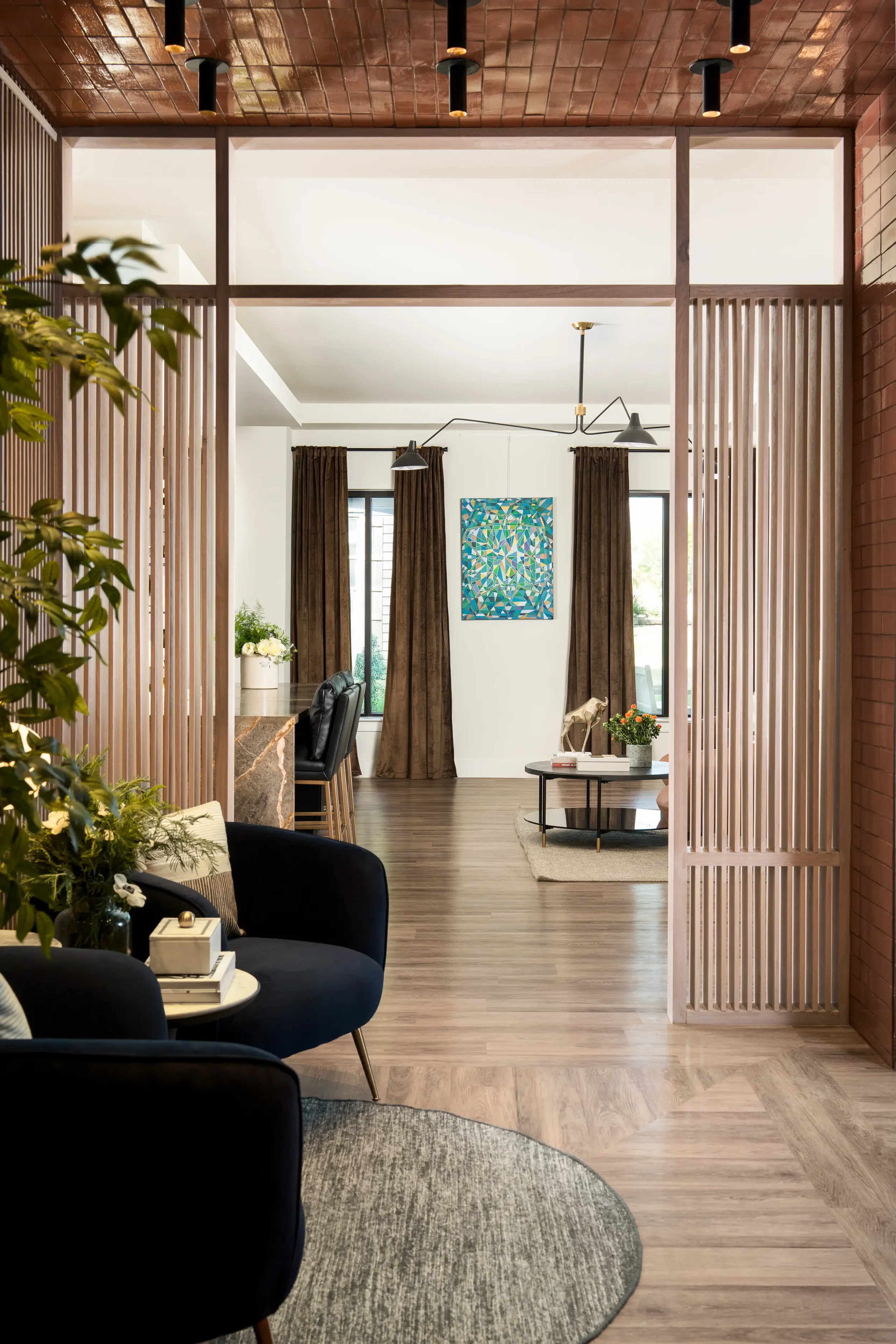 Interior view of a modern living room and dining area with wooden slat dividers, dark curtains, a colorful abstract painting, and a small round table with flowers and books. Designed by Artbuckle & Co. Located in Frisco, TX. 