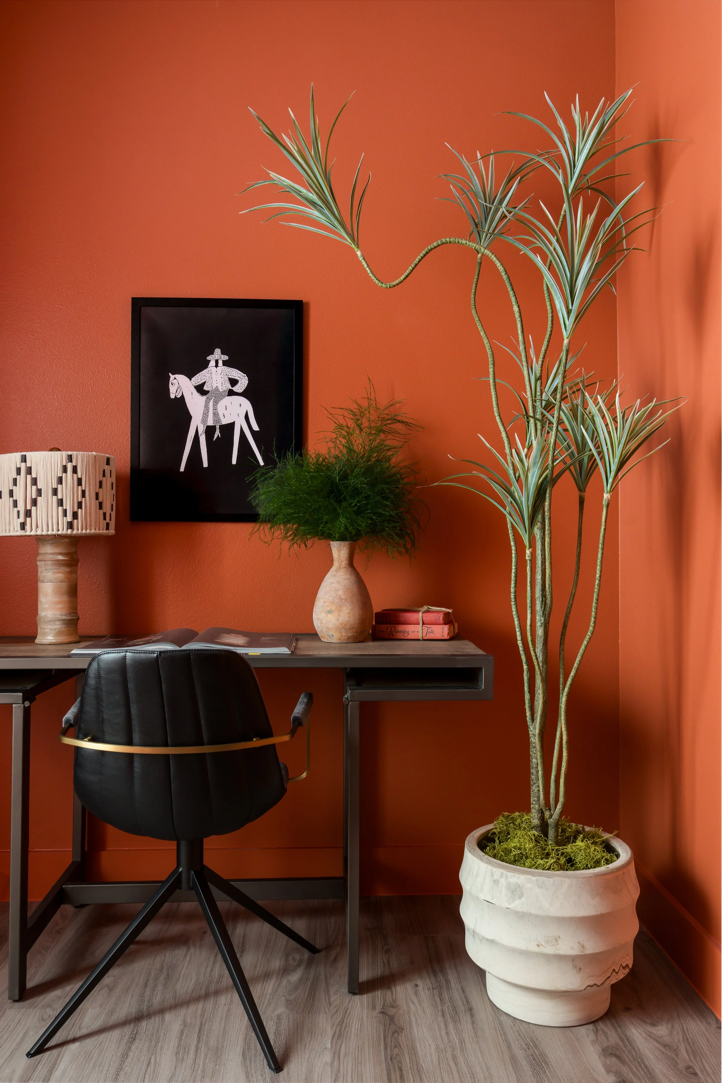 Home office with a black office chair in front of a desk against a burnt orange wall. On the wall, there's a framed black-and-white drawing of a man riding a horse. The desk has a tall potted plant with long, slender leaves and another, smaller plant