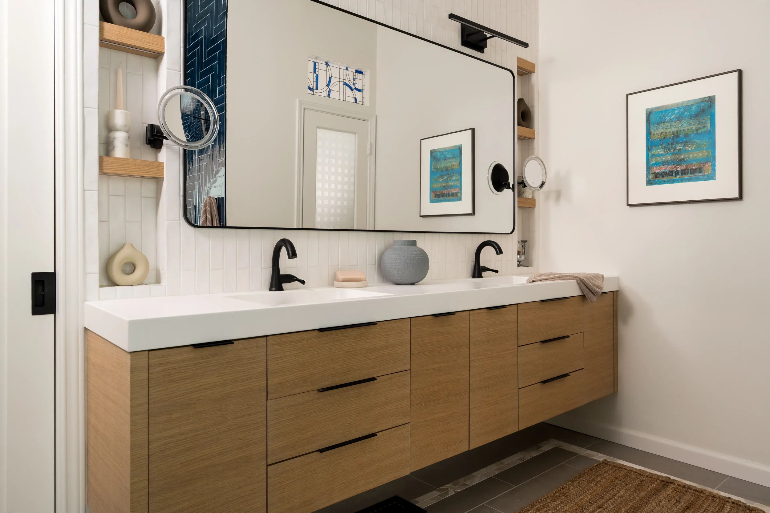 A modern bathroom vanity with a large rectangular mirror, two black faucets, wooden cabinetry, and wall-mounted wooden shelves decorated with vases. The wall has white shiplap paneling, and there are two framed abstract artworks hanging on the wall.