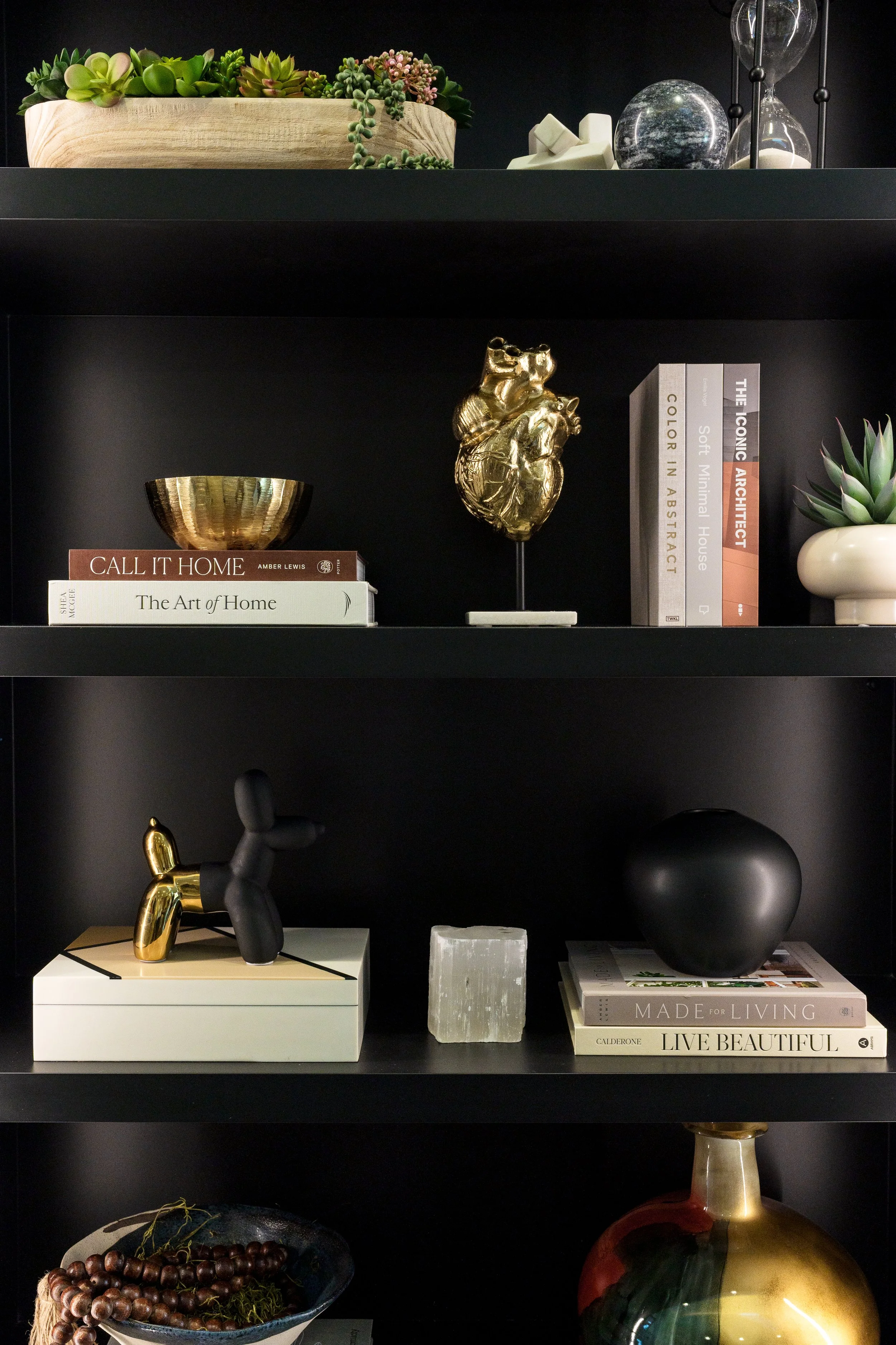 Black shelving unit displaying decorative objects and books: top shelf has succulents in wooden planter and glass orbs with sand; middle shelf has books about art and design, a gold anatomical heart sculpture, a plant in a white pot; lower shelf has 