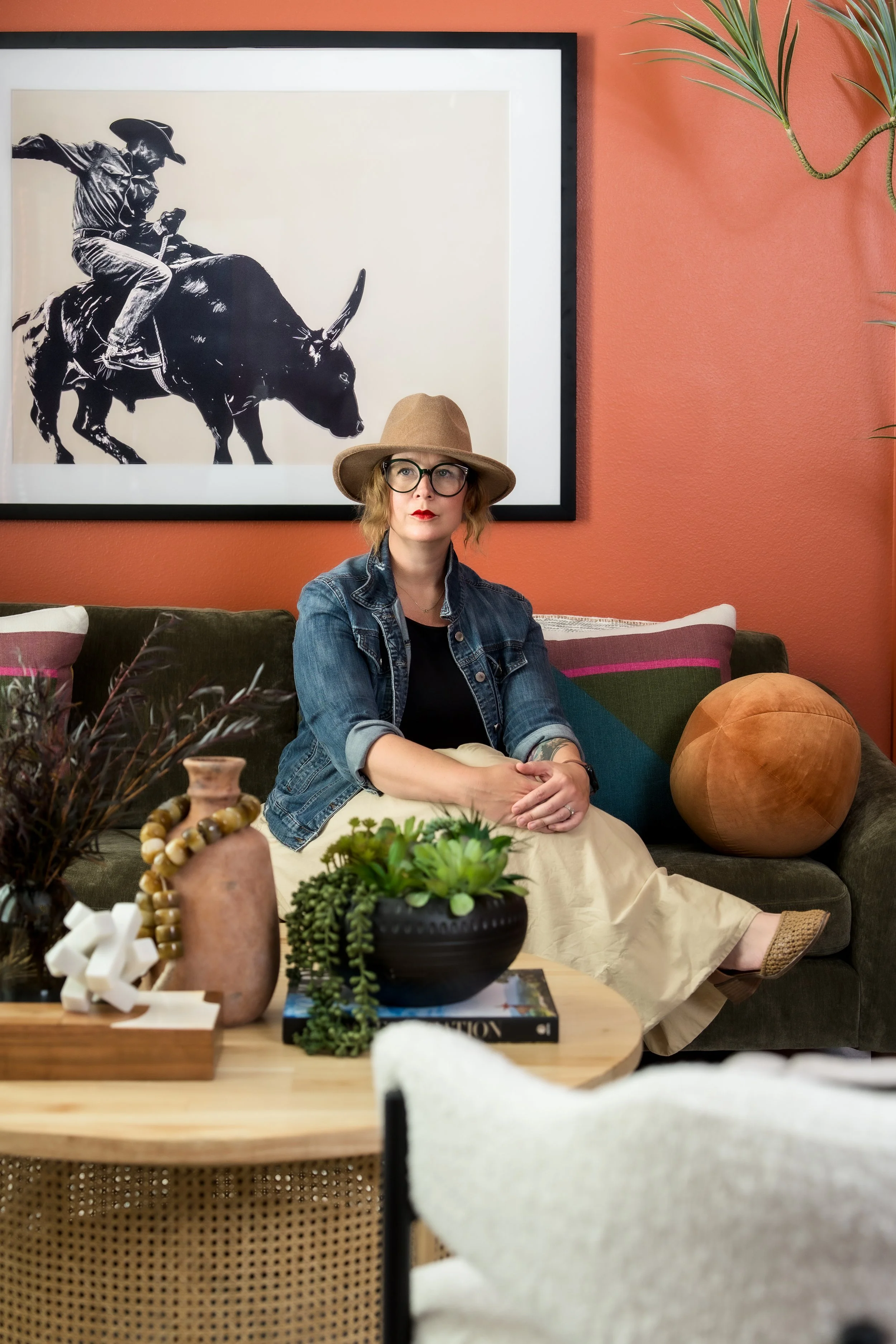 A woman with glasses, red lipstick, wearing a tan hat, a denim jacket, and a cream-colored skirt, sitting on a dark green sofa in a cozy room with pink walls. She is posing with her arms crossed and looking slightly to her right. Behind her is a fram