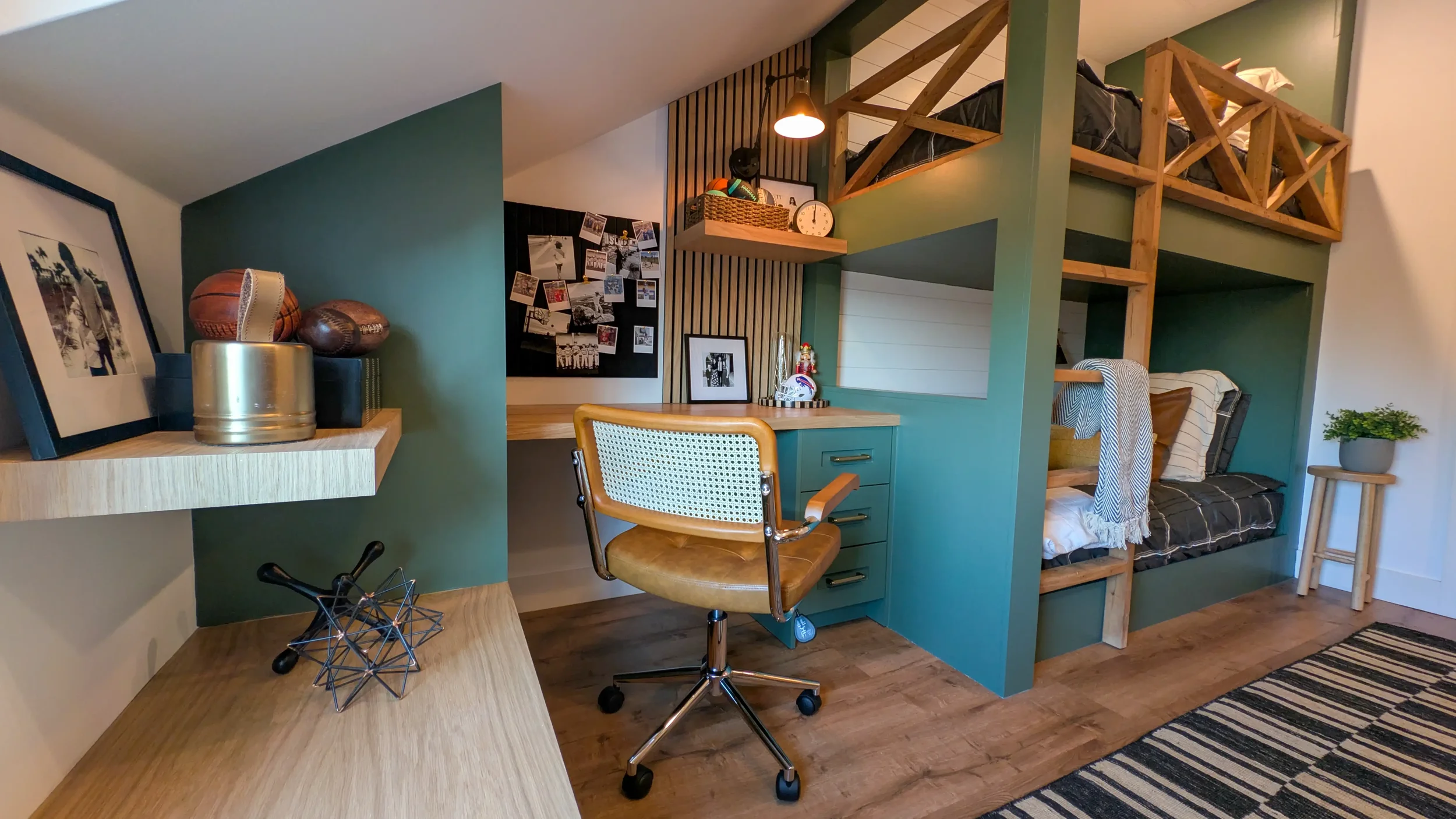 A cozy living space featuring a green accent wall with a wooden loft and bed above. There is a workspace with a desk and chair, decorated with framed photos, a clock, and a small Christmas ornament. A bench with pillows and a small round table with a potted plant are also visible.