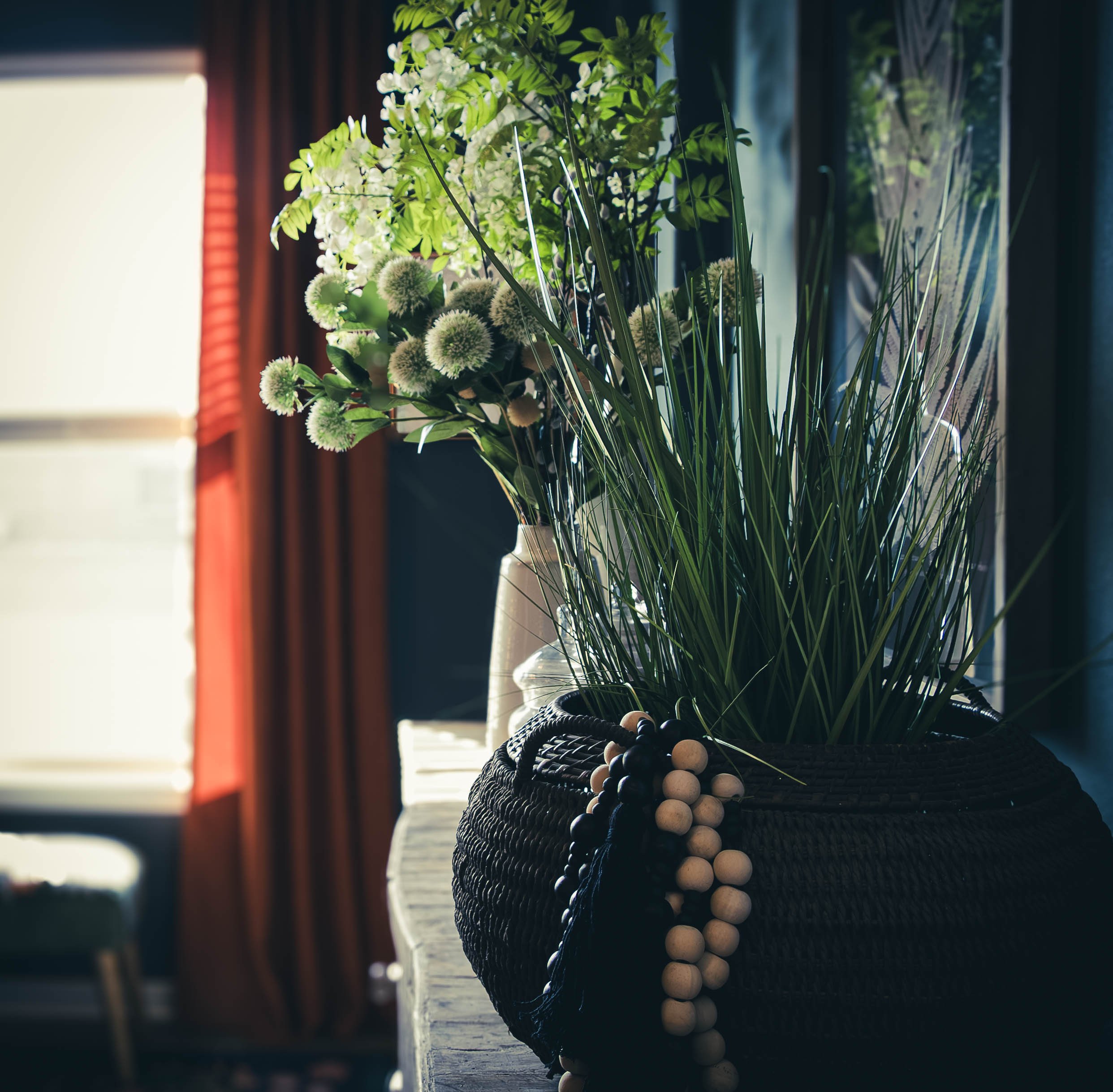A black woven basket filled with white and green flowers, including white round pom-pom-like blooms and green foliage, sitting on a wooden surface in natural sunlight, with a blurred background of a window and red curtains.