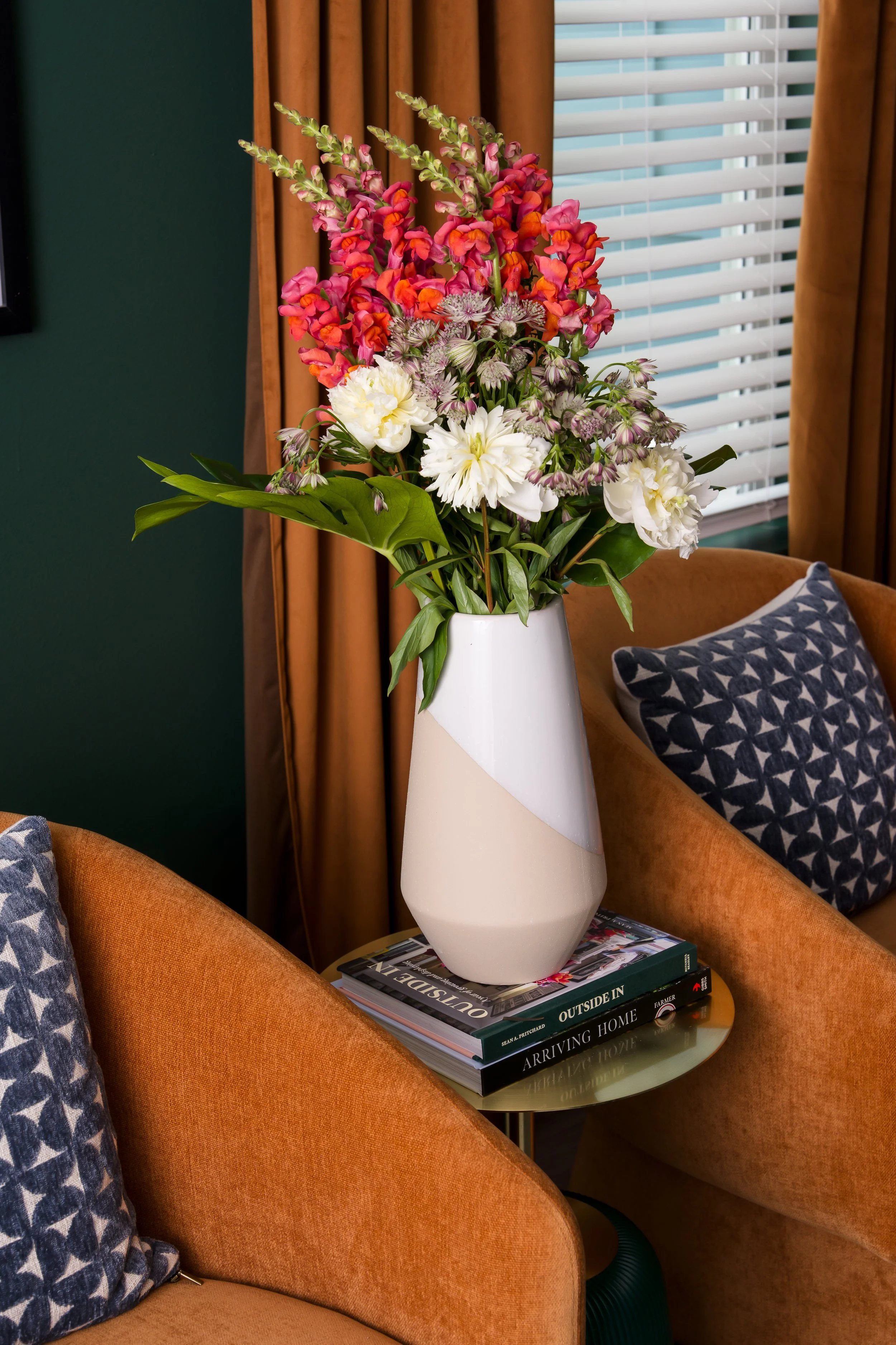 A tall white ceramic vase with a modern geometric design, filled with a colorful bouquet of pink, orange, white, and purple flowers, placed on a glass side table hosting a stack of several books.