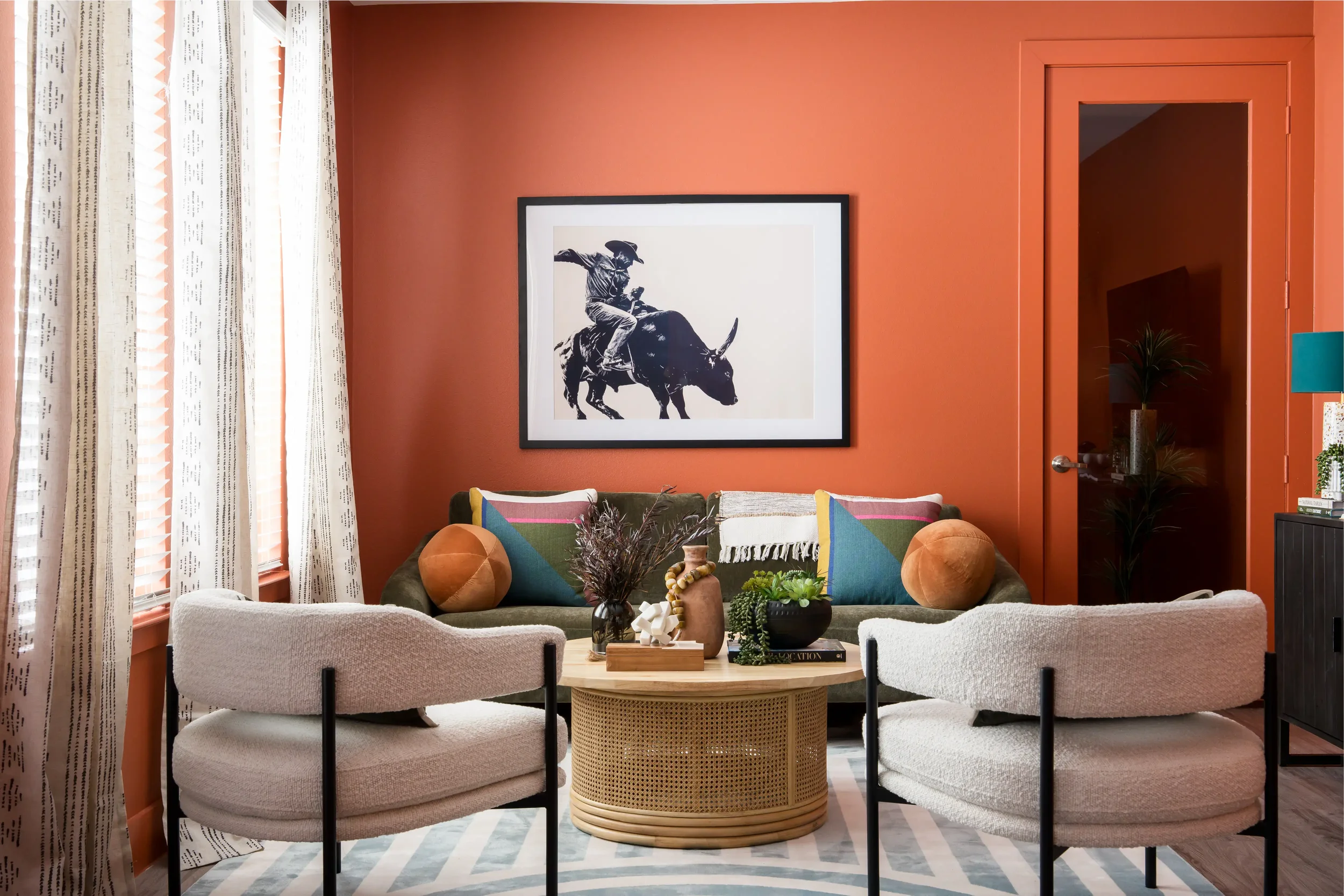 A living room with a coral-colored wall, a framed black and white print of a cowboy riding a buffalo, a white sofa with colorful pillows, a round wicker coffee table with decorative items, a beige armchair, window curtains with text patterns, and a g