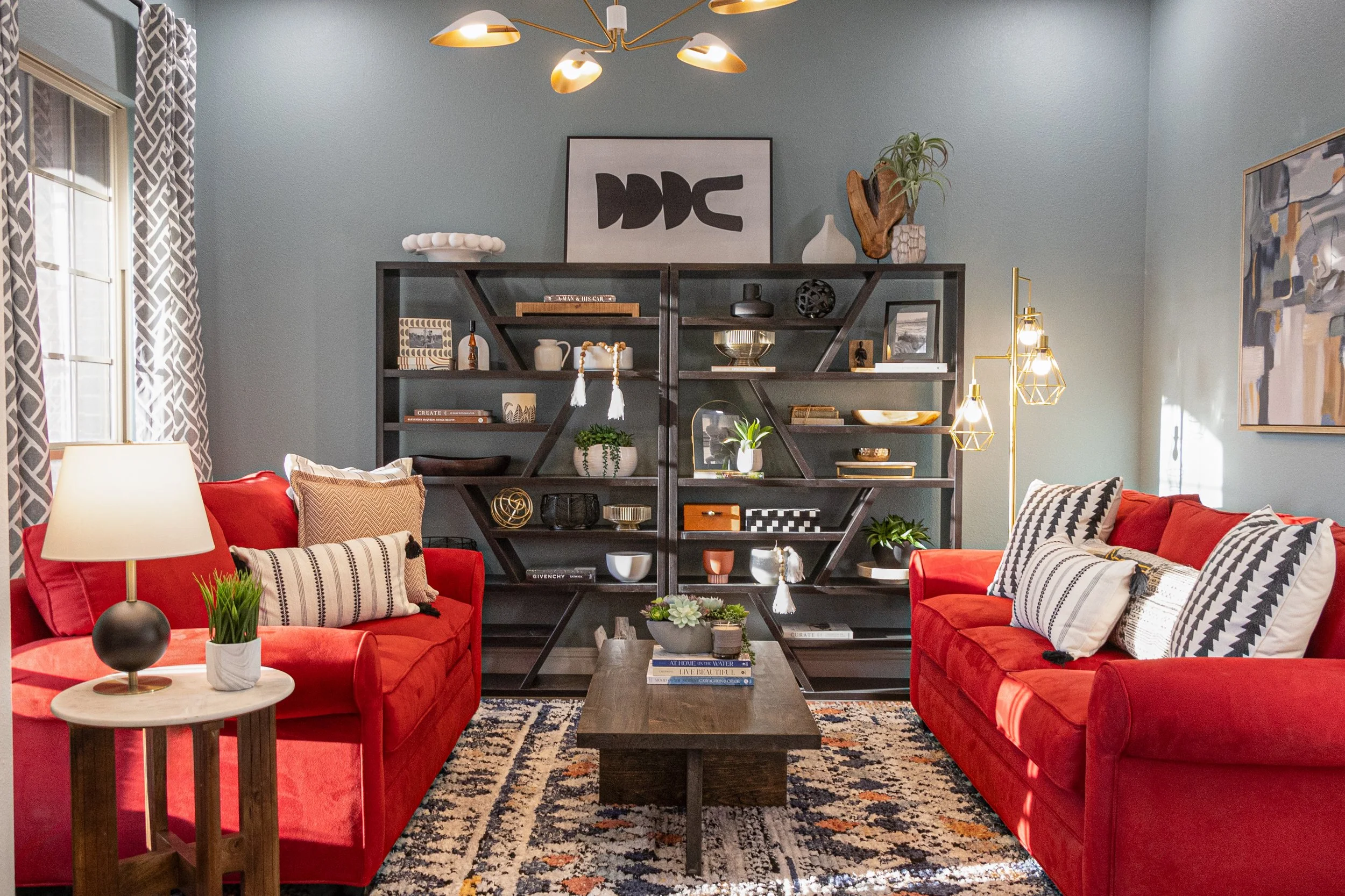 A cozy living room with two red sofas, patterned pillows, a wooden coffee table, a black bookshelf with decorative items, and a gold modern chandelier. There are two windows with patterned curtains, and a large abstract painting on the wall.