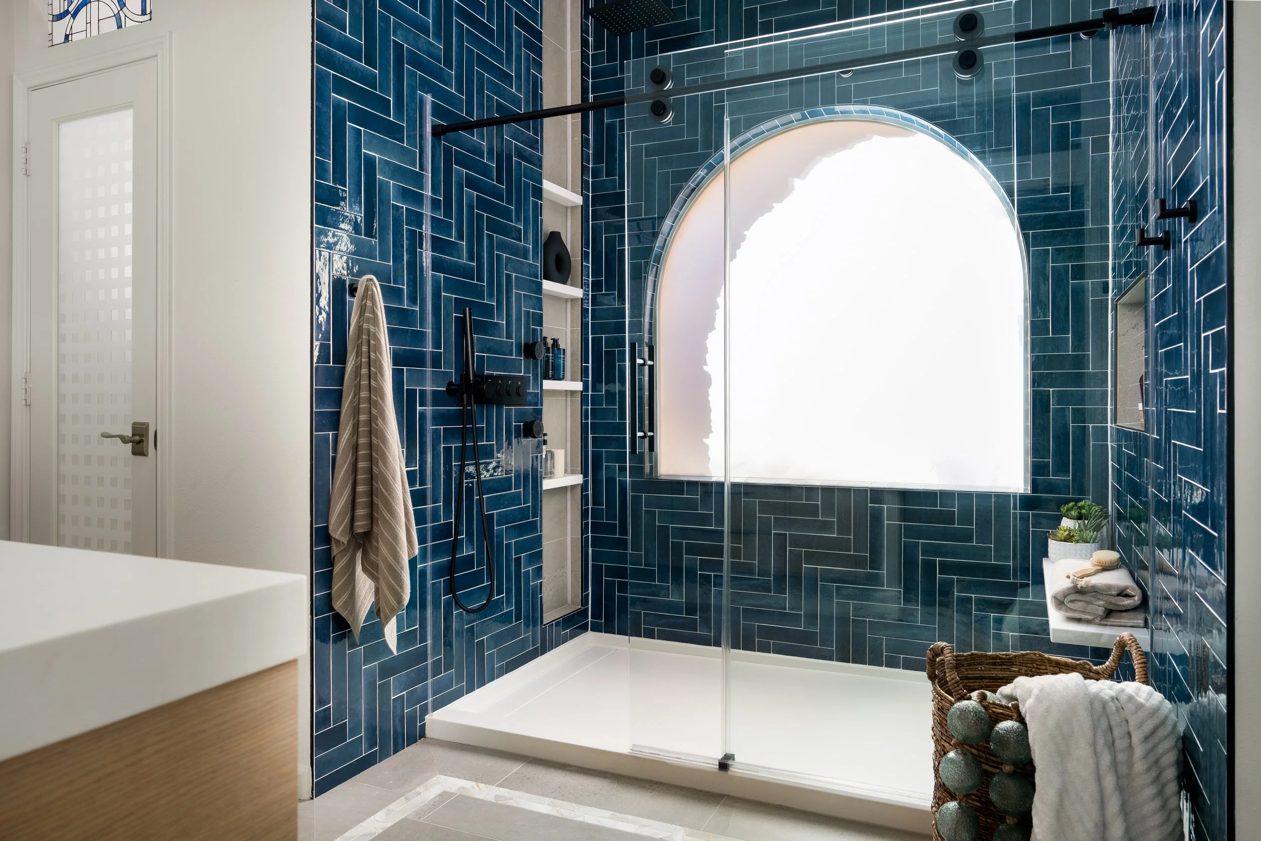 Modern bathroom with dark blue tile walls surrounding a glass-enclosed shower area with a large arched window. The shower has black fixtures, a built-in shelf, and a black showerhead. There are towels and decor items on a small shelf and in a woven b