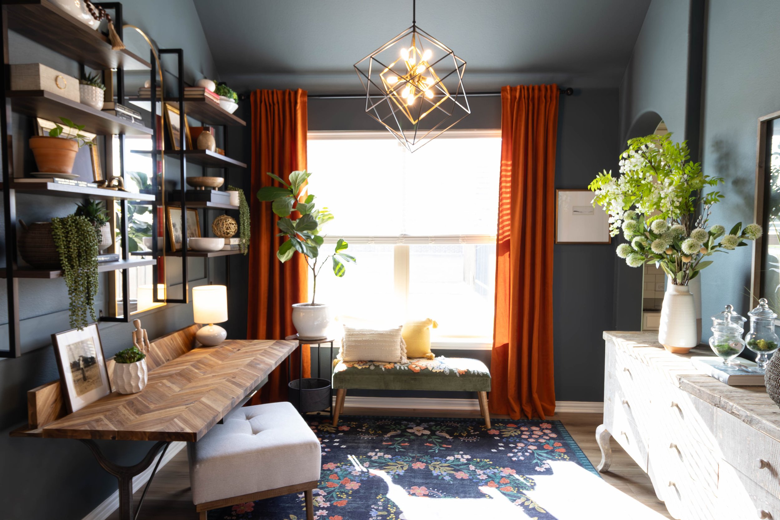 A cozy living room with a dark accent wall, bookshelves with decorative items, a window with orange curtains, a modern geometric chandelier, and a floral area rug with furniture and houseplants.