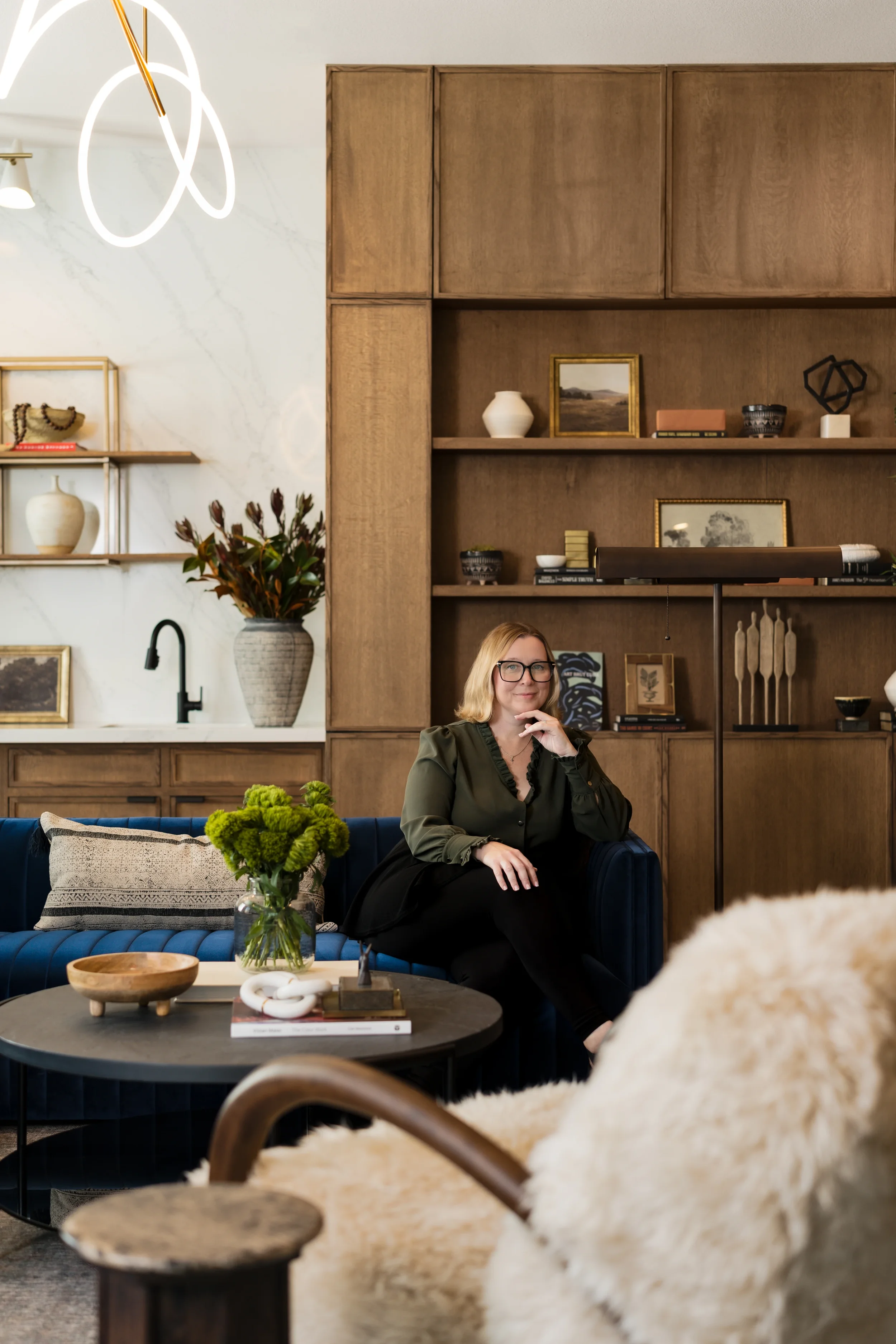 Rebecca Gillispie of Artbuckle & Co. sitting in the completed luxury resident lounge at The Gallery in Frisco, TX, featuring custom walnut millwork, blue velvet seating, white shearling chair, and modern loop chandelier.