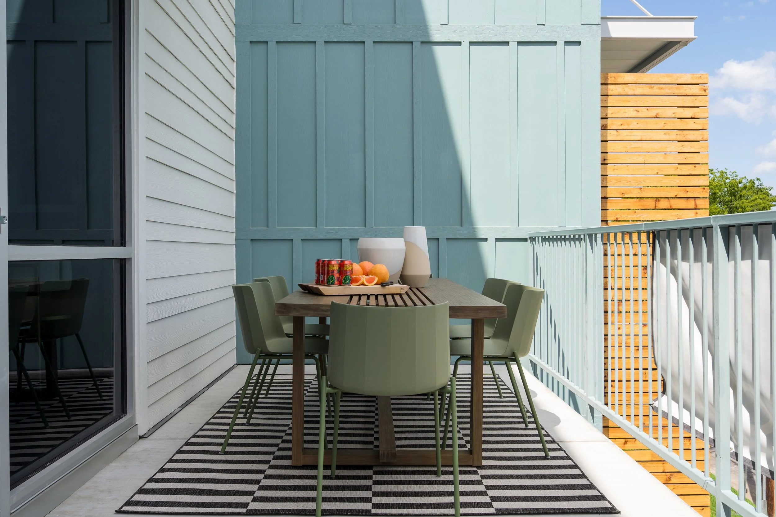 Outdoor balcony with a wooden dining table, six green chairs, a striped black-and-white rug, and a tray of drinks, fruits, and white vases. The balcony has white railings, light blue and white exterior walls, and a wooden privacy screen at the far en