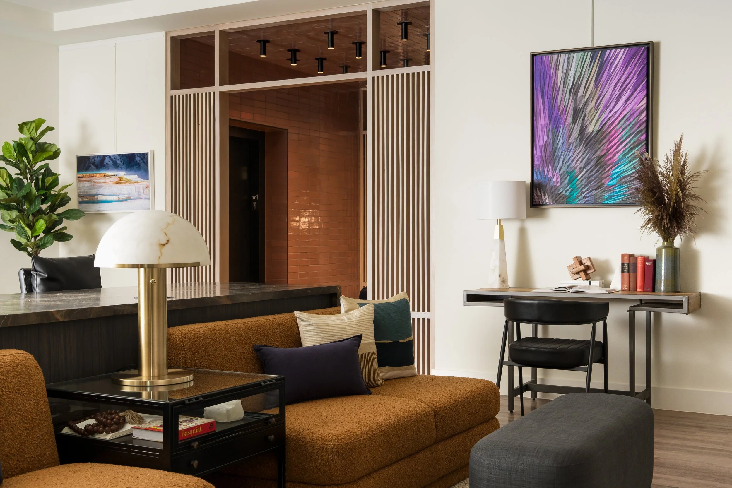 Living room interior with brown couch and colorful pillows, black coffee table with books and decorative items, large plants, modern white wall with artwork and a large colorful digital screen, wood accent ceiling, and a small workspace with a desk, 